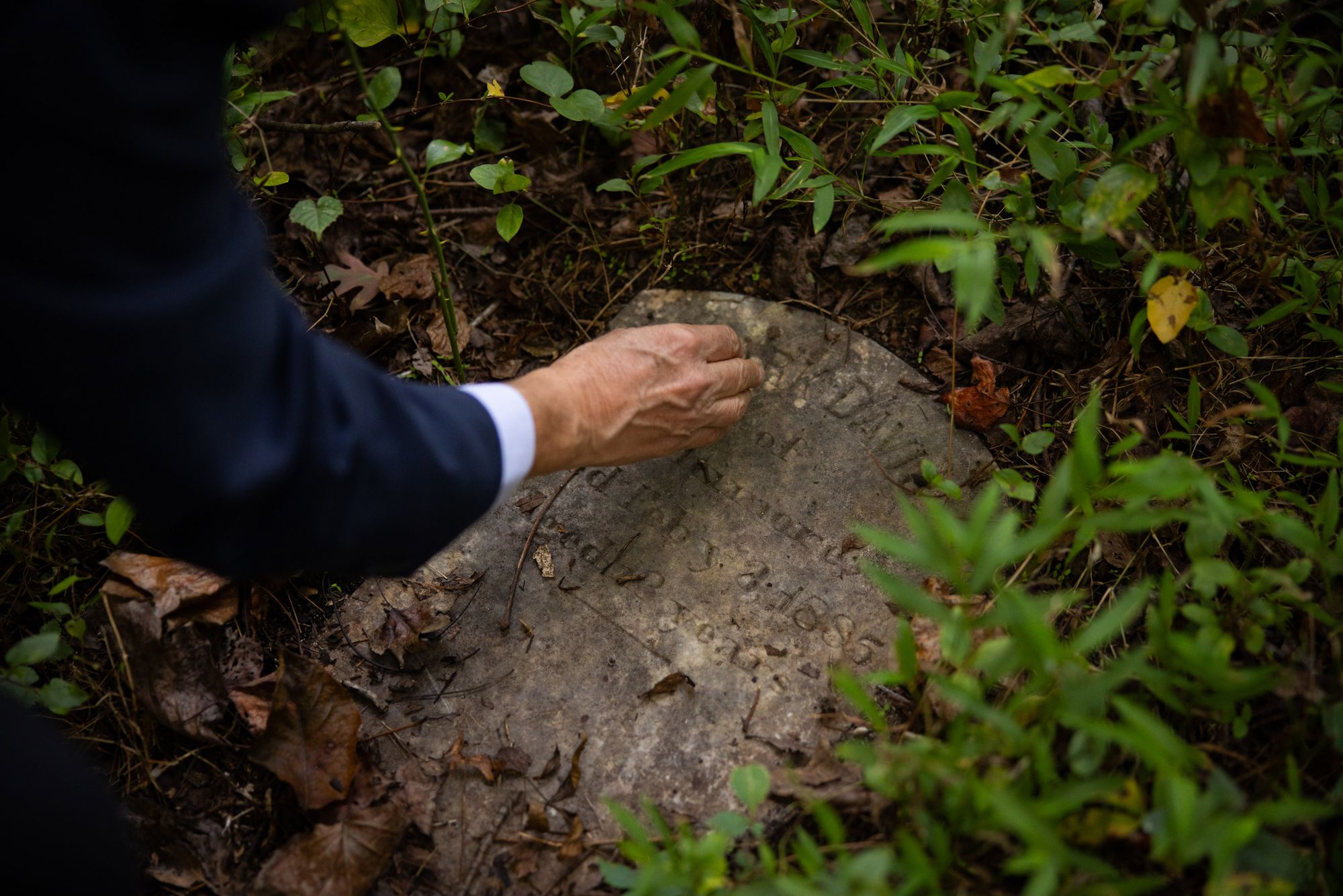 A close-up photo of a White man's hand moving debris off a gravestone covered in dirt and surrounded by plants on a forest floor.