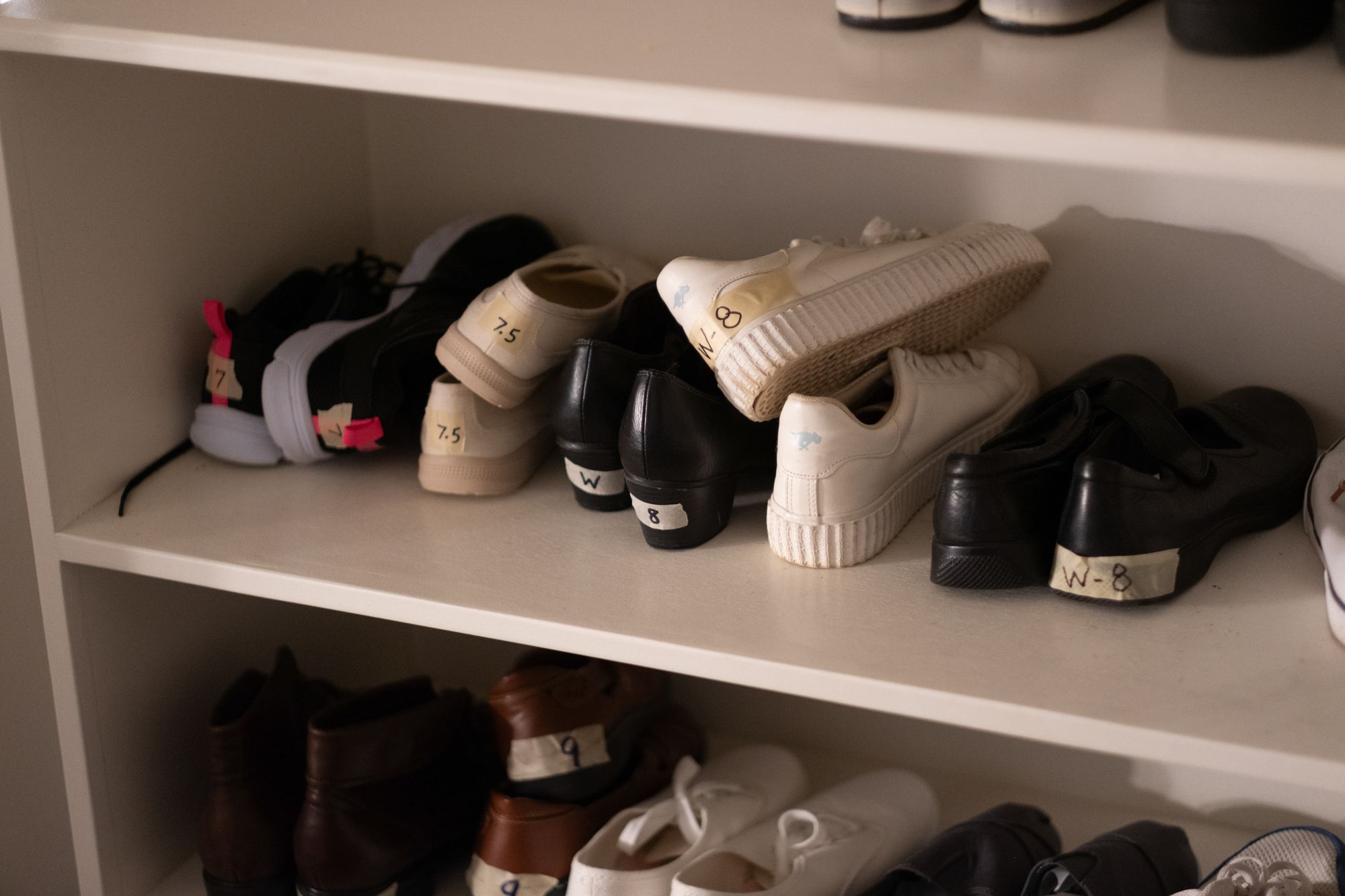 Eleven pairs of shoes, including sneakers and leather shoes in white, black, beige and brown, are lined up on white shelves.