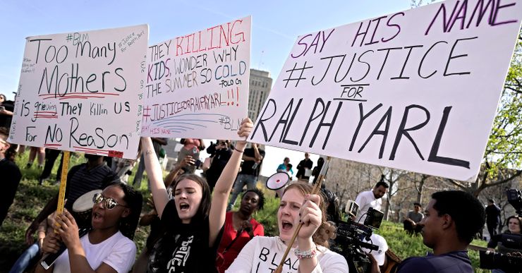 Protesters at a park hold signs that reads "Too many mothers is grieving, they killing us for no reason," "They killing kids, wondering why the summer so cold. Justice for Ralph Yarl!!" and "Say his name, justice for Ralph Yarl."  People with bullhorns and cameras stand in the background. 

