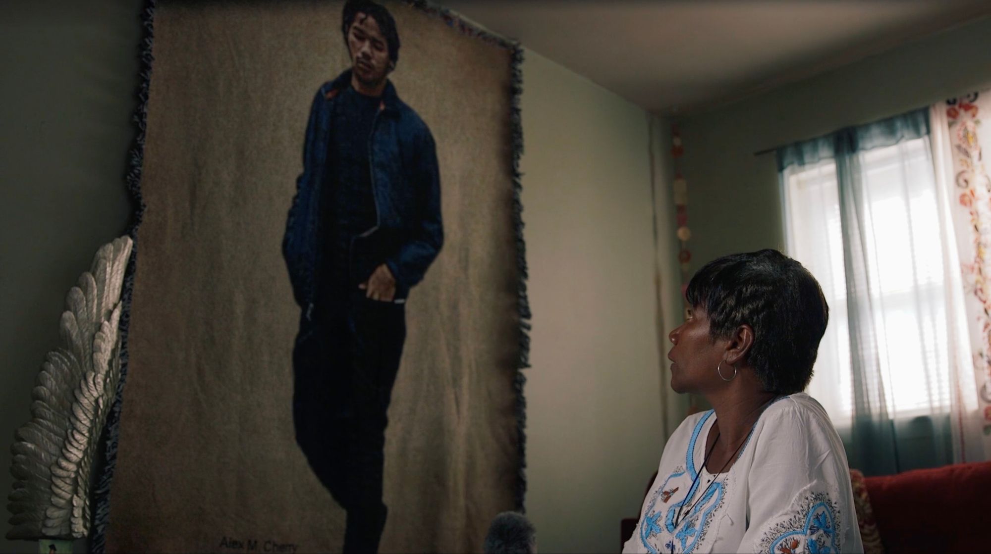 Christina Cherry, a woman with dark brown skin and sitting on a couch in her home, looks upward at a blanket hanging on the wall. The blanket depicts an image of her son Alex, who was shot and killed in 2016. 