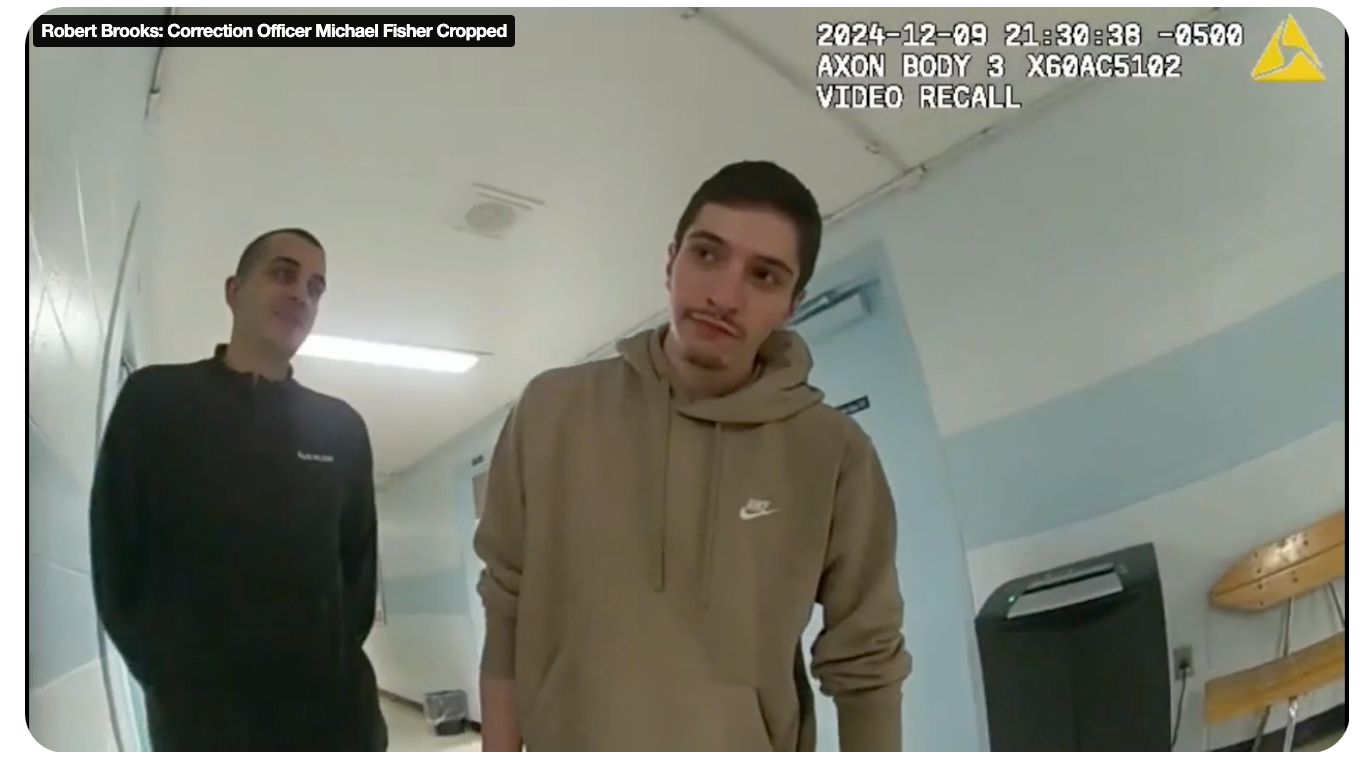 A man in a black jacket, on the left, and a man wearing a tan hooded sweatshirt stand in a light blue hallway. 