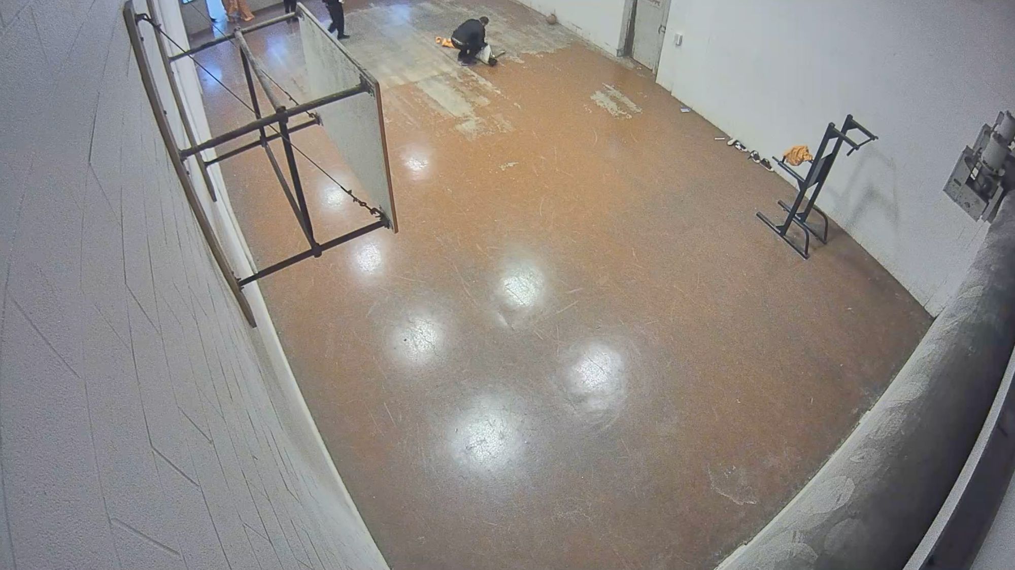 Three still images from a surveillance camera show an indoor gym at a jail. In the first frame, a man in an orange and white jail uniform is on the ground as other men approach him. In the second frame, a correctional officer kneels over the man on the floor. In the third frame, a group of correctional officers surround the man on the floor.