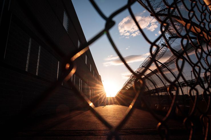 A photo shows a sun setting in between a building and barb wired fences. Dusk lighting make the building appear as a silhouette. In the foreground is a close-up of a wired fence. 