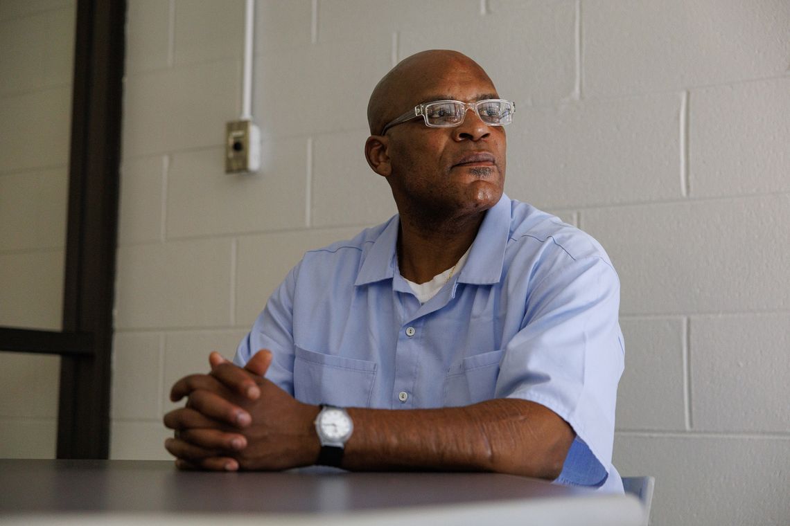 A photo shows a Black man, wearing glasses, a watch and a blue prison uniform. 