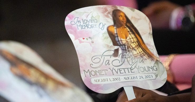 The hand of a Black person holds a fan of an image of a Black woman wearing a black and white plaid dress. The fan reads: "In loving memory of Ta'Kiya Monet Yvette Young. August 1, 2002 - August 24, 2023." 