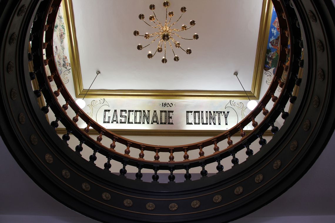 A photo looking up through a circular wooden railing shows the ceiling of a courthouse. There are paintings, golden trim molding, and text that reads “Gasconade County” on the ceiling.