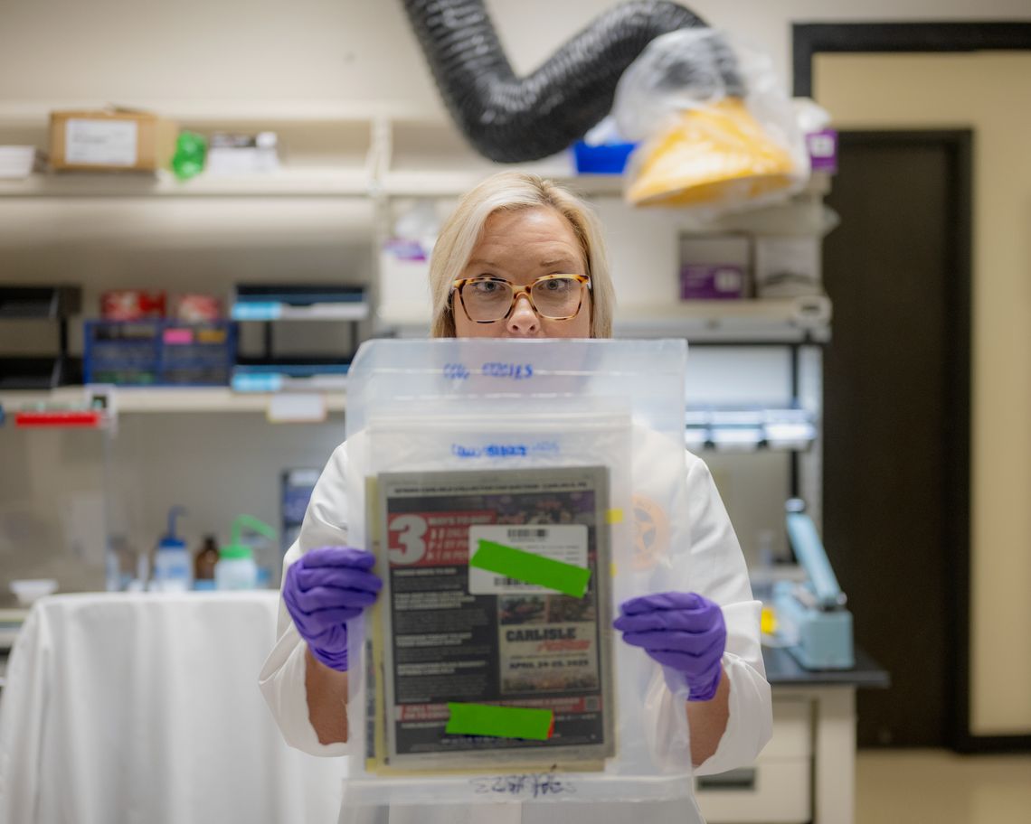 A photo shows a White woman with blonde hair wearing a lab coat and purple gloves holding up a plastic bag that has some papers and folders in it.