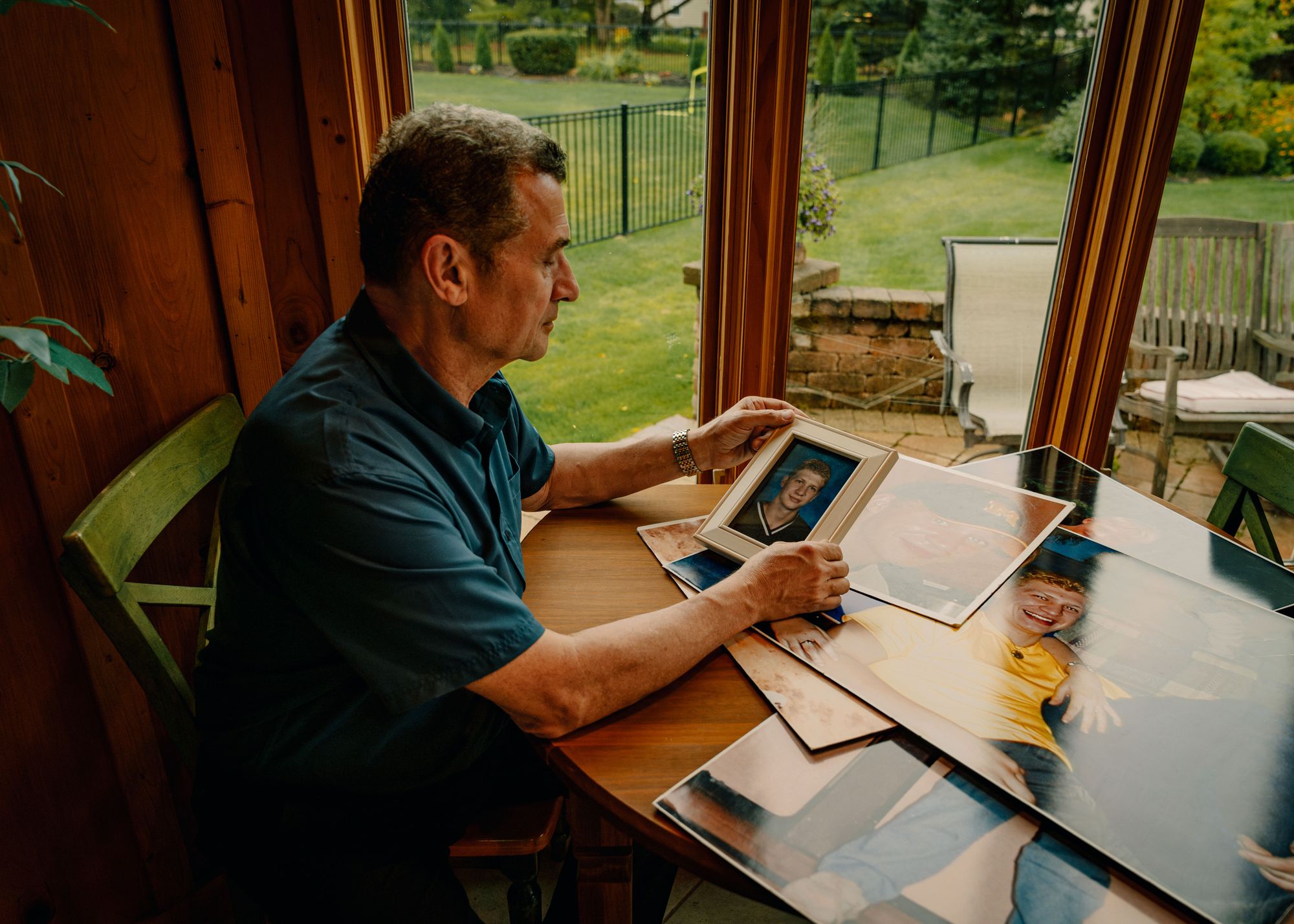 Michael Bell Sr., a White man, looks at a high school photo of his son, Michael Bell Jr., a White man, while sitting at a table. Four other photos of Michael are visible beneath the high school photo of Michael Bell Jr.