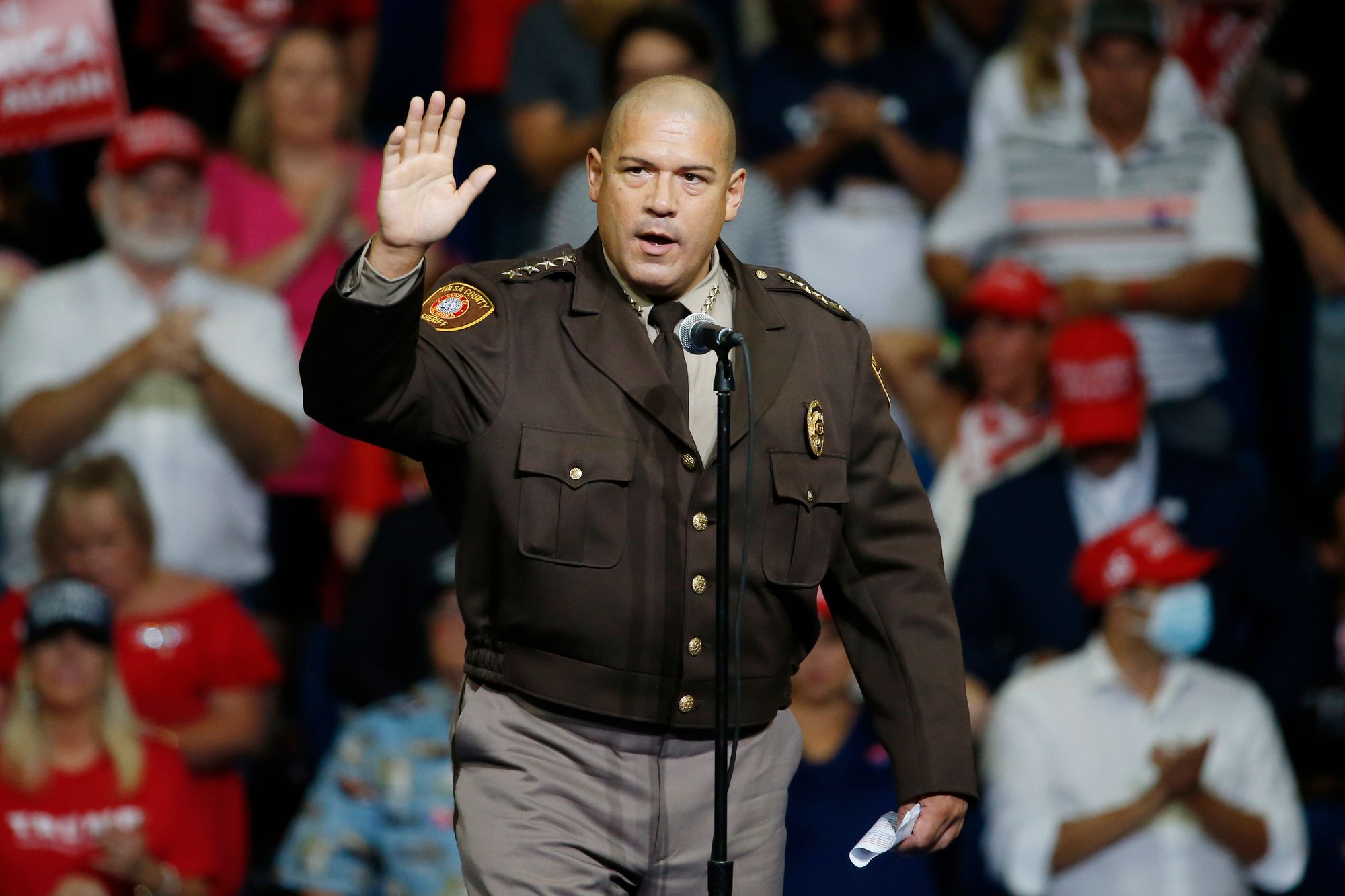 On the left, a White man with a shaved head and dressed in a brown sheriff’s uniform and jacket, stands at a microphone at a political rally and raises his right hand to the crowd.