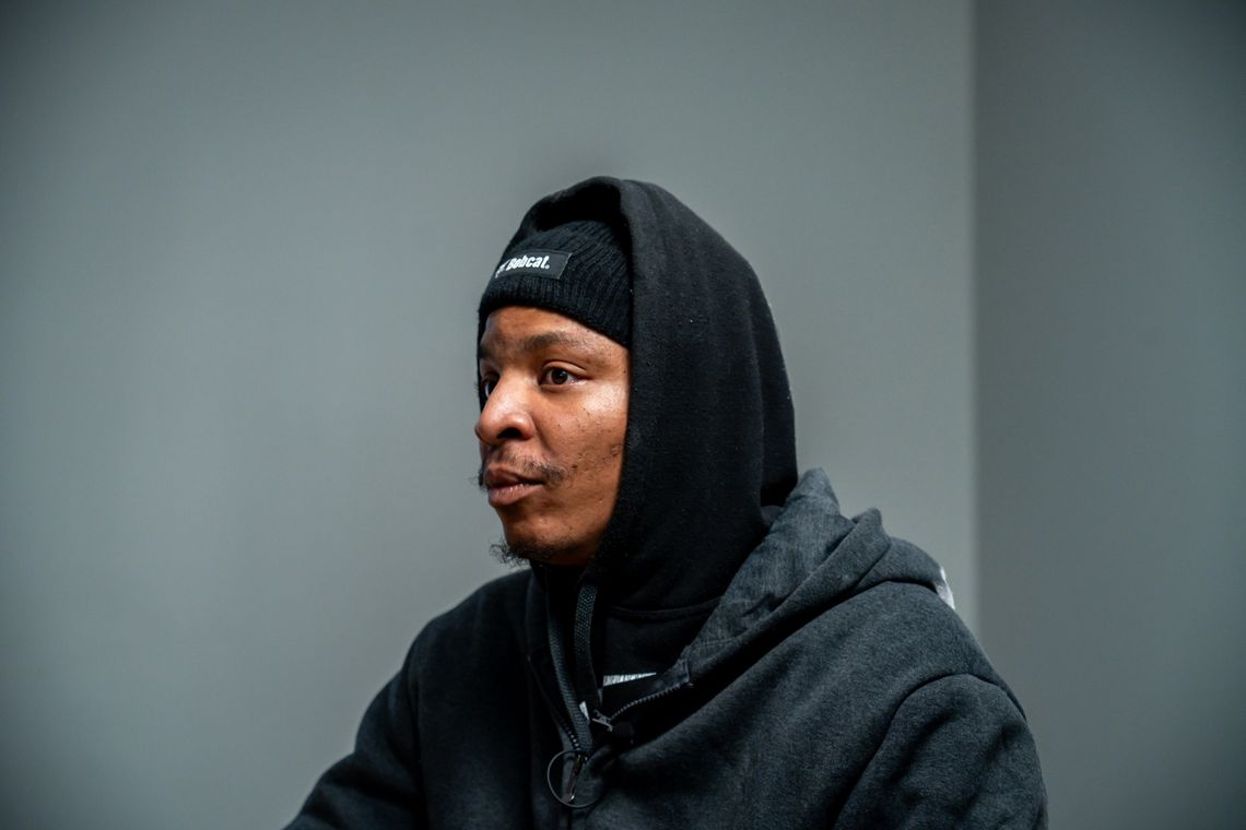 A photo shows the head and shoulders of Tim Wade, a Black man wearing a black hoodie sweatshirt, jacket, and beanie, as he looks to the left. 