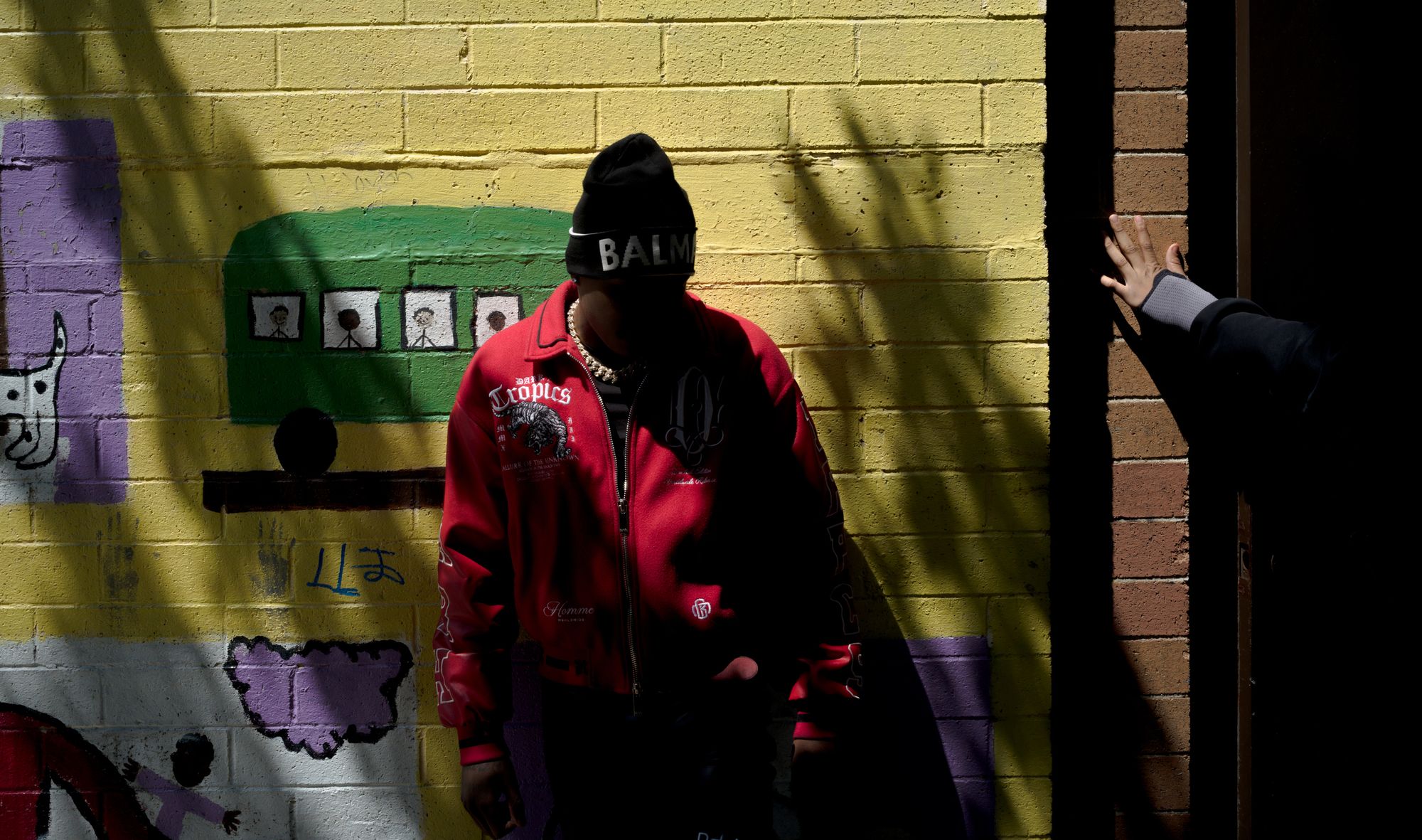 T-Man, a Black teen wearing a red coat, stands in front of a yellow wall that has a mural painted on it. His face is not visible.  