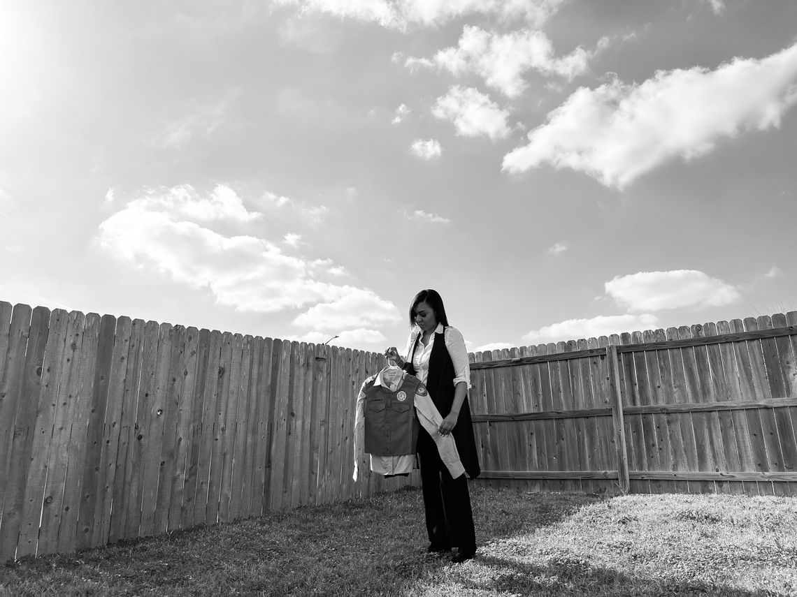 A black-and-white photo shows a Black woman with medium-length hair, a white long-sleeved shirt and a black vest holding a uniform. Behind her is wooden fencing, and the sky with clouds is visible.