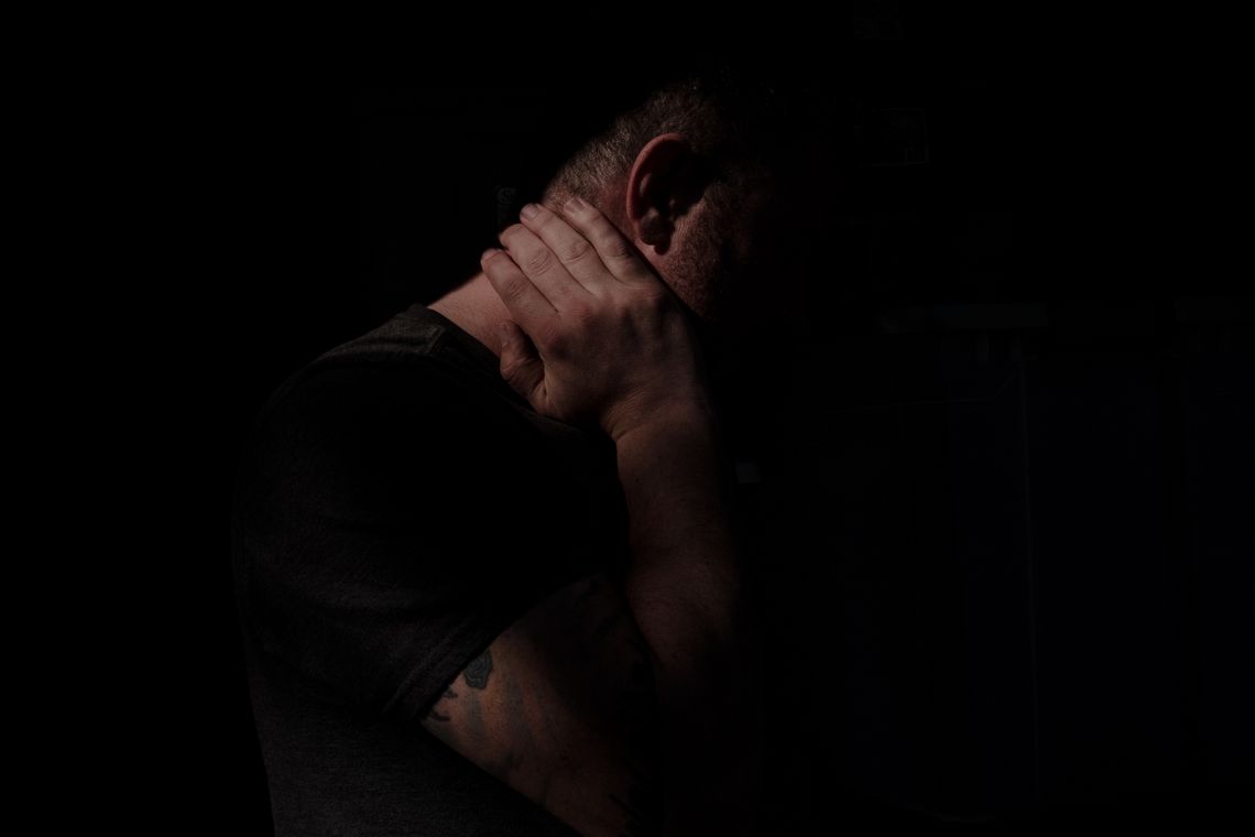 A White man with close-cut dark hair stands in shadow, his right hand resting on his neck below his right ear. 