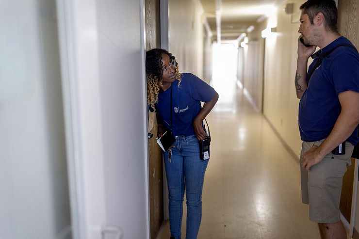 A Black woman, wearing glasses, a blue T-shirt and jeans, leans against a door as she holds a notebook and a walkie talkie. She looks at her colleague, a White man, wearing a blue T-shirt and khaki shorts, as he is talking to a cell phone. 