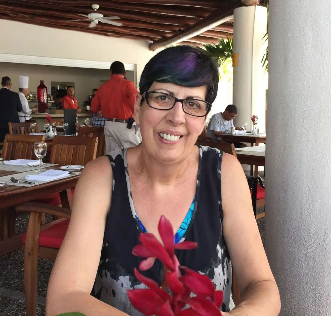 A White woman with light-toned skin and short, dark brown hair, wearing glasses, smiles at the camera.