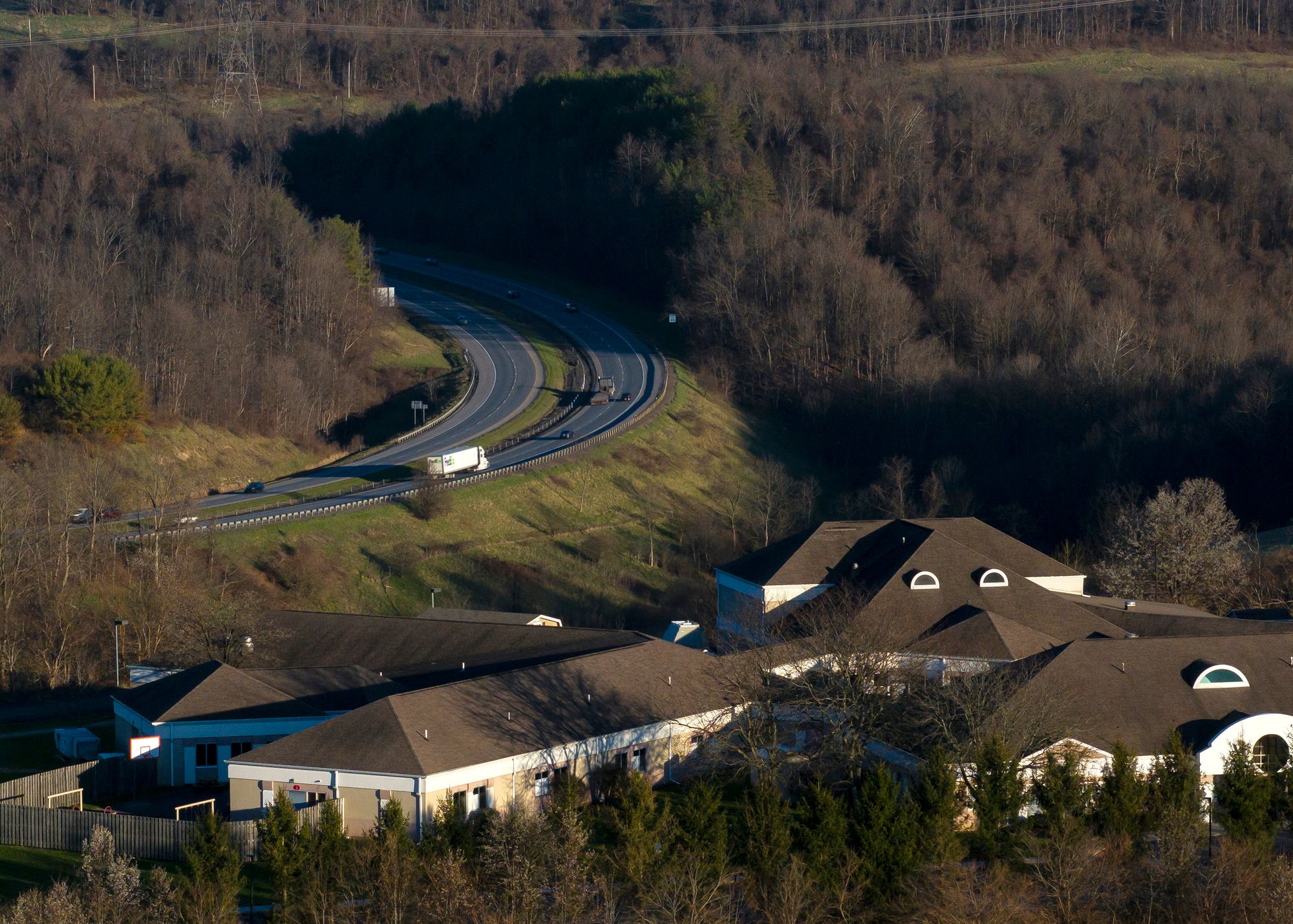 A photo shows a low beige-colored building with a dark roof at the bottom of the image. Above and to the left is a highway with some vehicles driving on it. Trees surround the highway and buildings.
