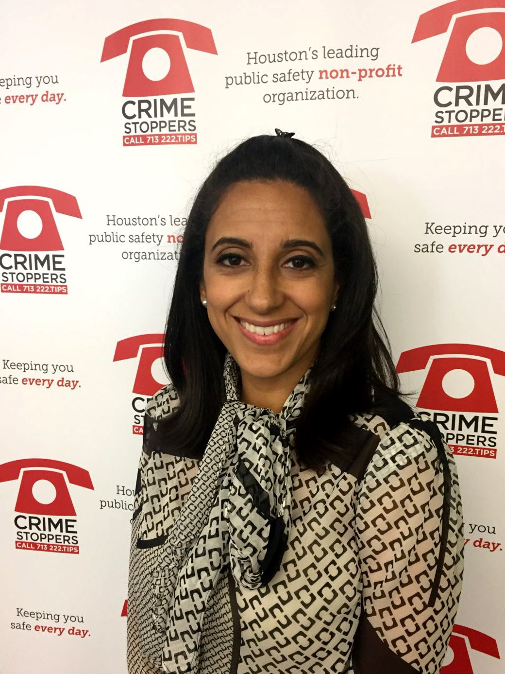 A woman with medium-toned skin and long dark hair wears a black and white blouse in front of a back drop that has the Crime Stoppers logo on it. 