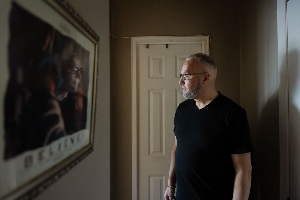 A photo shows a Latif Hafroui, a Moroccan man, standing in front of a door at the end of a hallway. He is looking to the left in the photo. A framed poster is visible on the left side wall with the word “Believe” on it. 