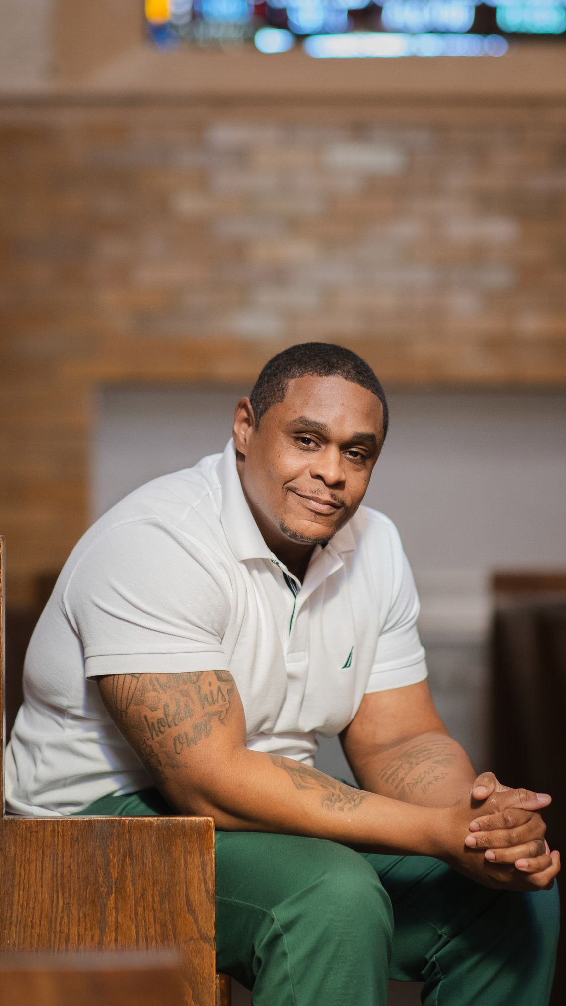 A Black man with a slight smile and tattooed arms sits in a church pew, wearing a light-colored polo shirt and green prison pants. He leans forward with his elbows on his knees, his hands clasped. 