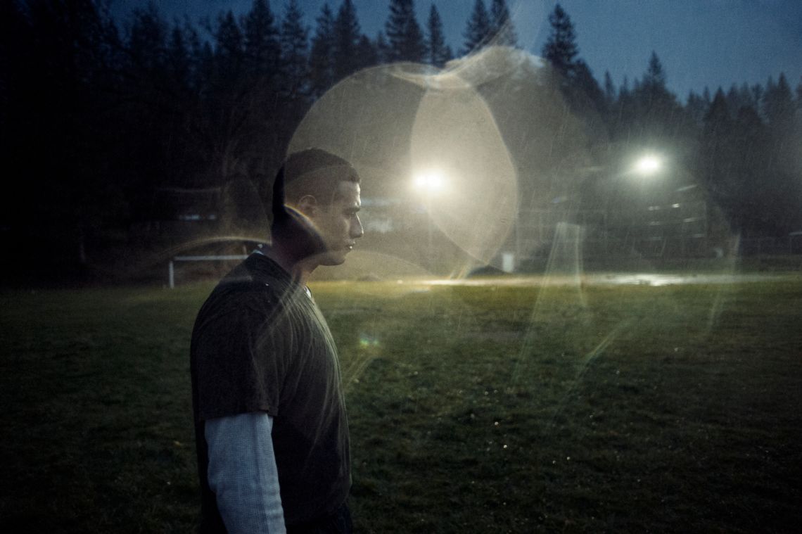 A young man with short, dark hair and wearing a T-shirt over a long-sleeved shirt stands on a green sports field at dusk.  Bokeh from light shining through drops of water are visible over part of the image.  Trees are visible in the background.   
