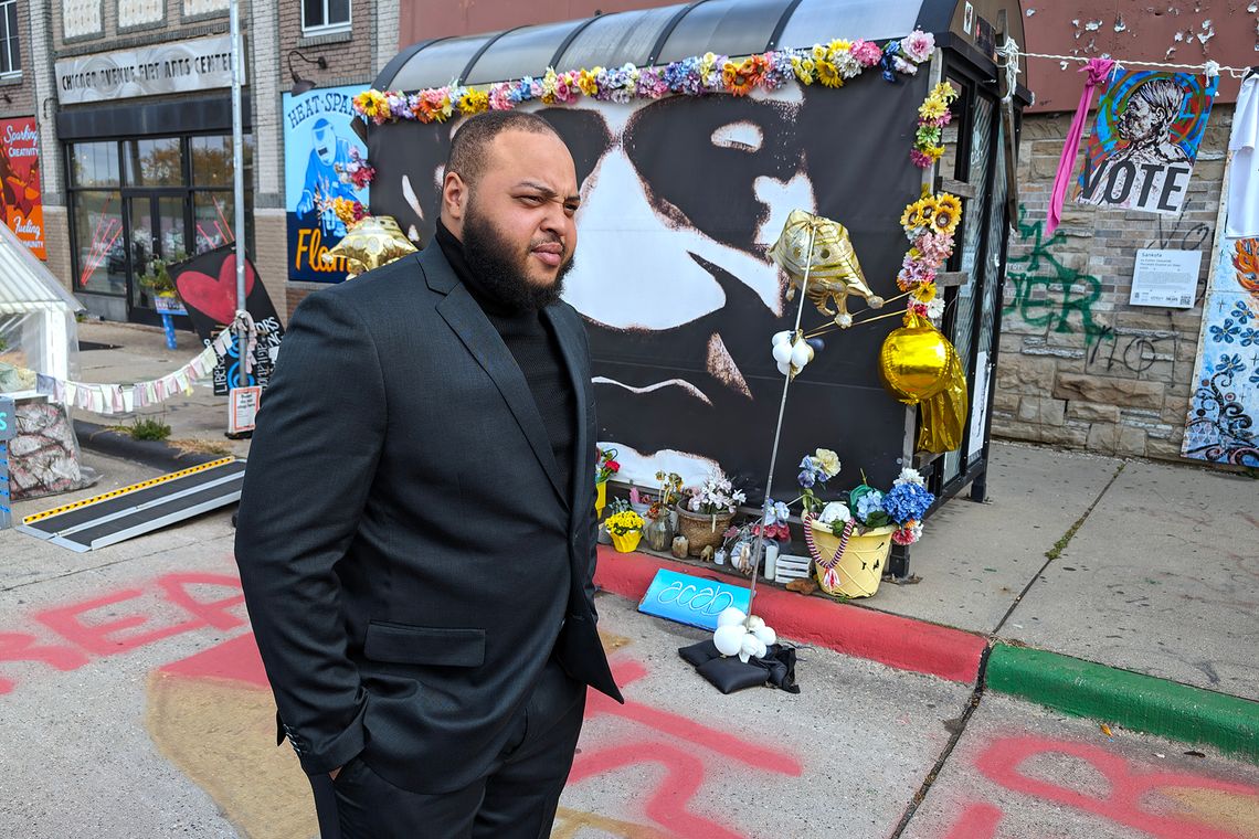 A Black man, wearing a black suit, looks pensive while standing in front of a memorial on a street. 