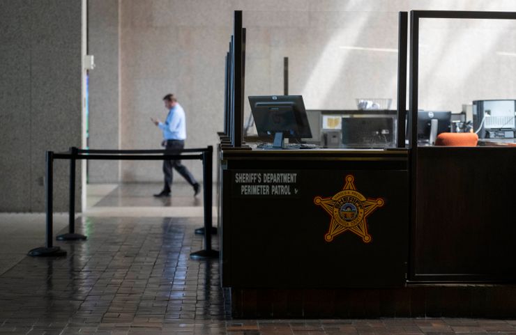 A sectioned off area inside the Cuyahoga County Justice Complex labeled “Sheriff’s Department Perimeter Patrol” has computers and chairs.  A man in a long-sleeved button-down shirt and dark-colored pants walks towards the left, behind the sectioned off area.  