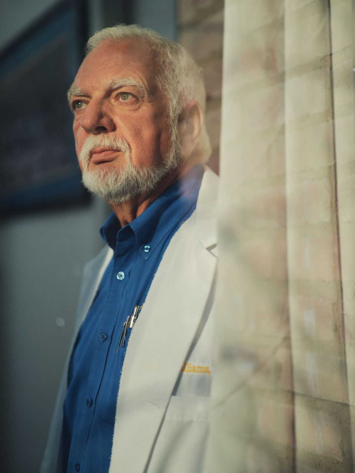 An older White man with white hair and a beard, wearing a white medical coat and blue shirt, stands in front of a window.