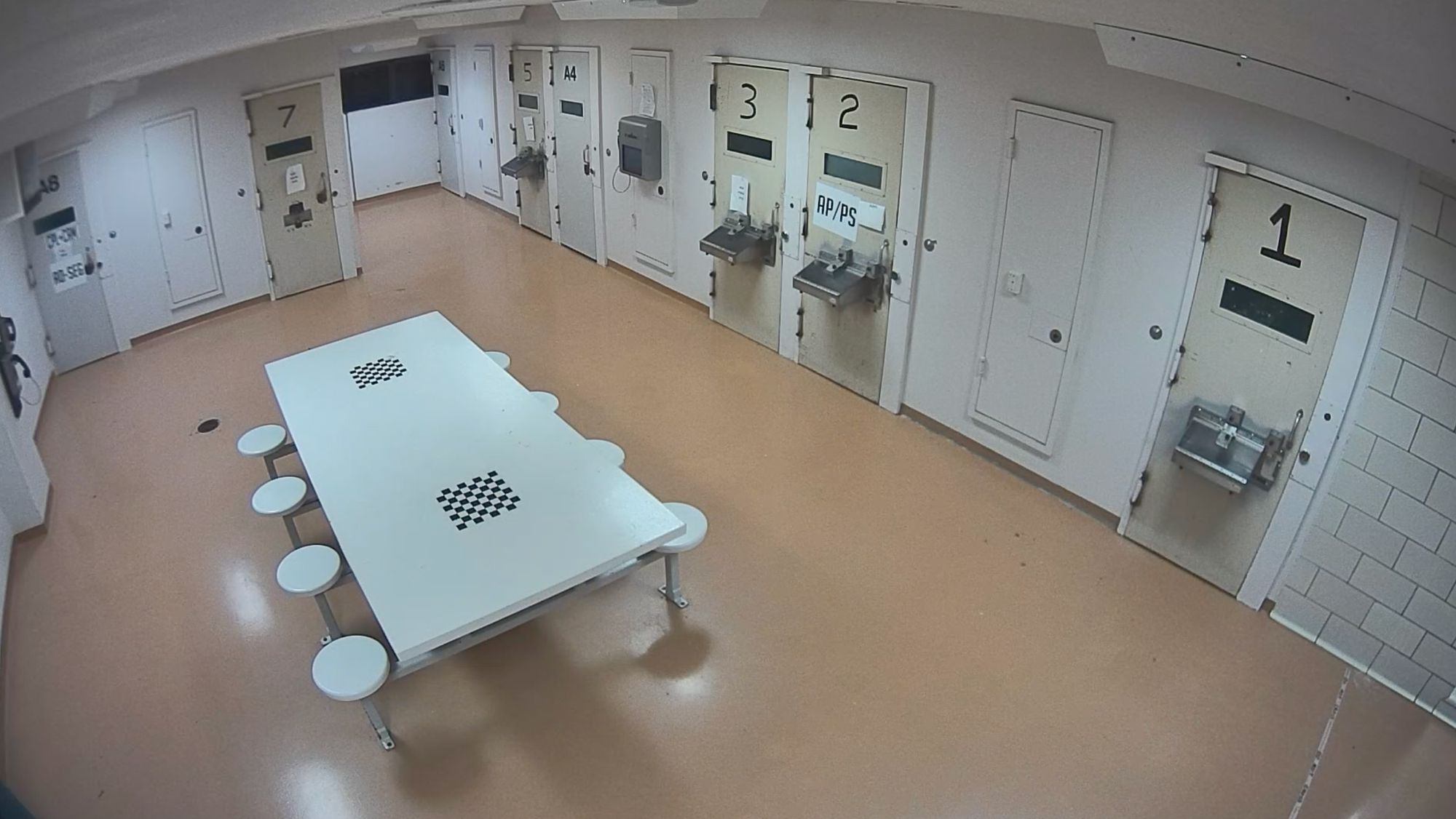 Two still images from a surveillance camera show a pod of jail cells. In the first image, the pod is empty. In the second image, two correctional officers are standing outside a door with the number “7.” 