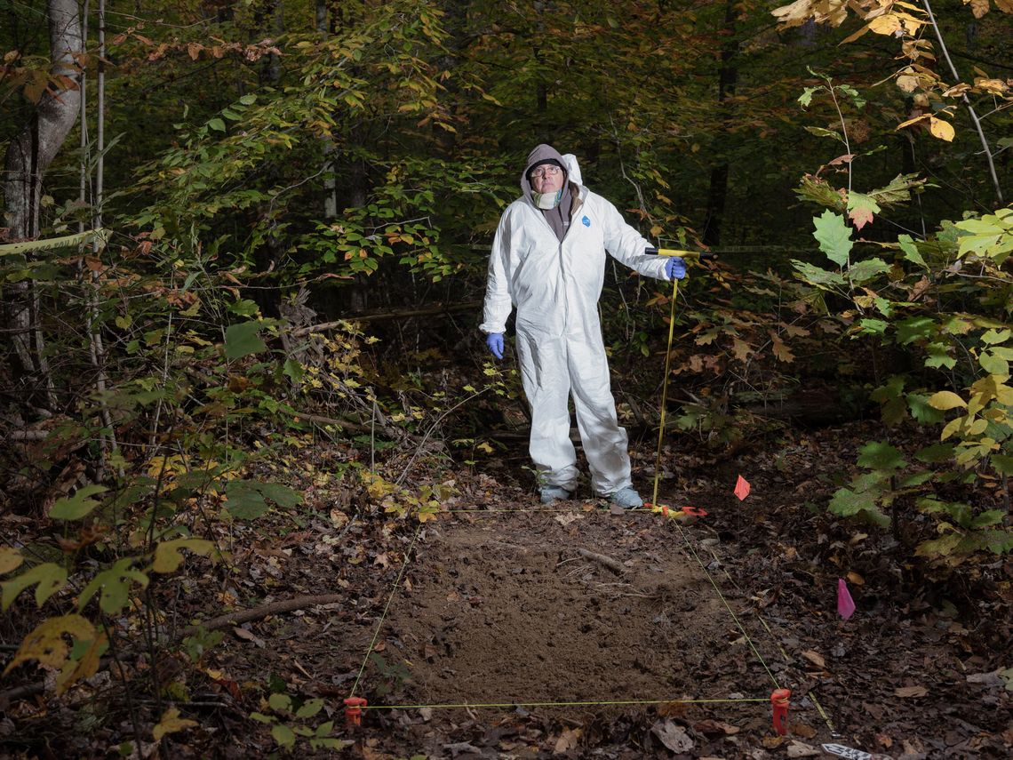 Arpad Vass, a Caucasian man wearing glasses and a white hazmat suit, stands in the woods in front of a unmarked grave delineated by yellow string in a rectangle shape. 