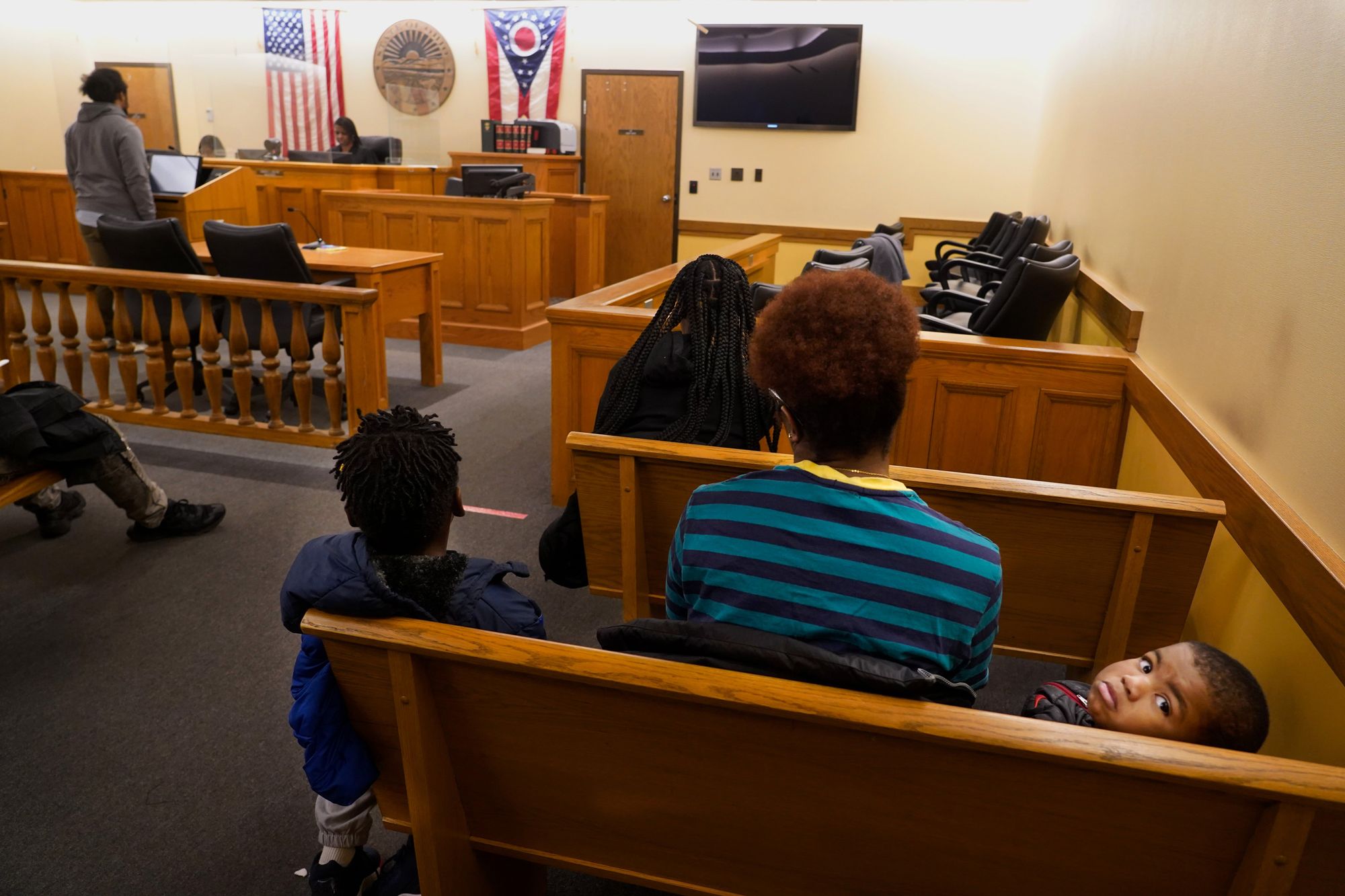 A view from the back of a courtroom, as a Black woman judge hears a case. In the back row, a Black woman wearing a blue and black striped shirt sits with two children.