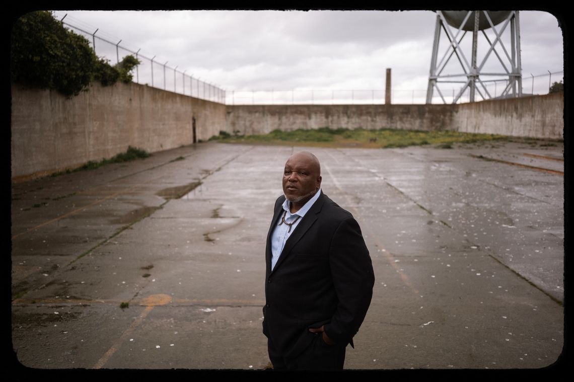 A photo shows Troy Williams, a Black man wearing a black suit and a light blue shirt, standing outdoors in a space surrounded by concrete walls. The ground has some puddles on it from rain. 