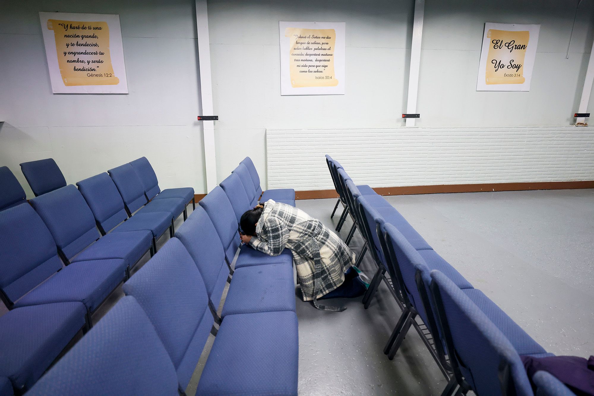 A woman, wearing a black and white plaid jacket, kneels in between a row of blue chairs. Three signs of Bible verses translated into Spanish hang on the wall. 