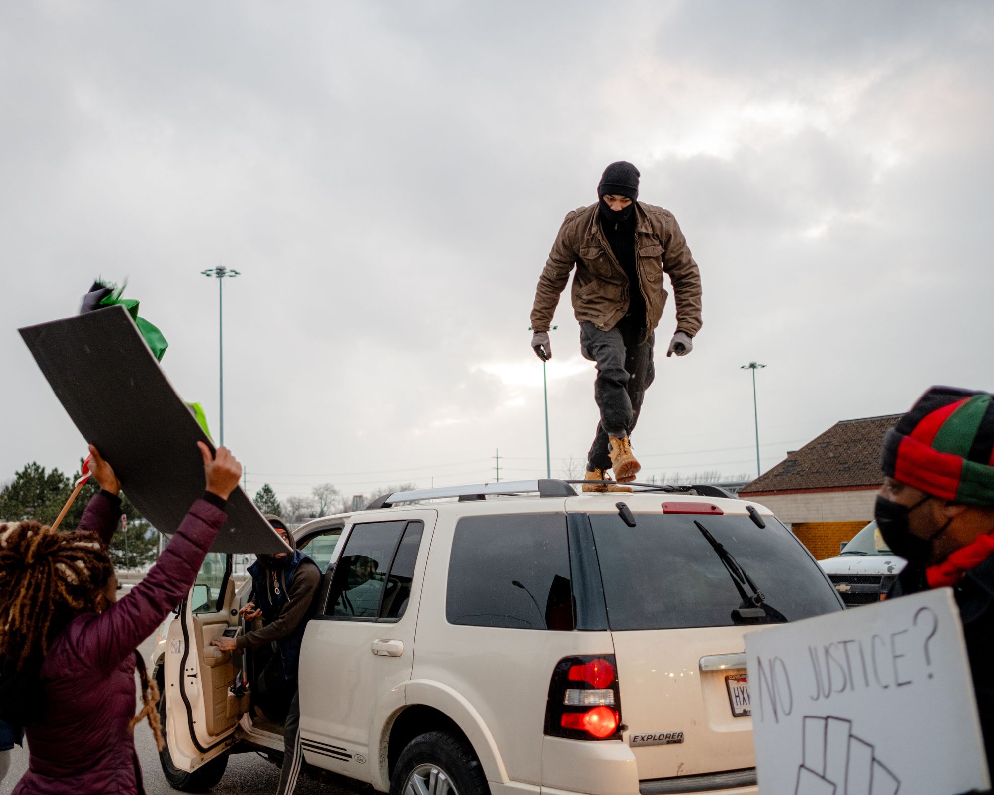 A photo of a masked person standing on top of a white van during a protest. 