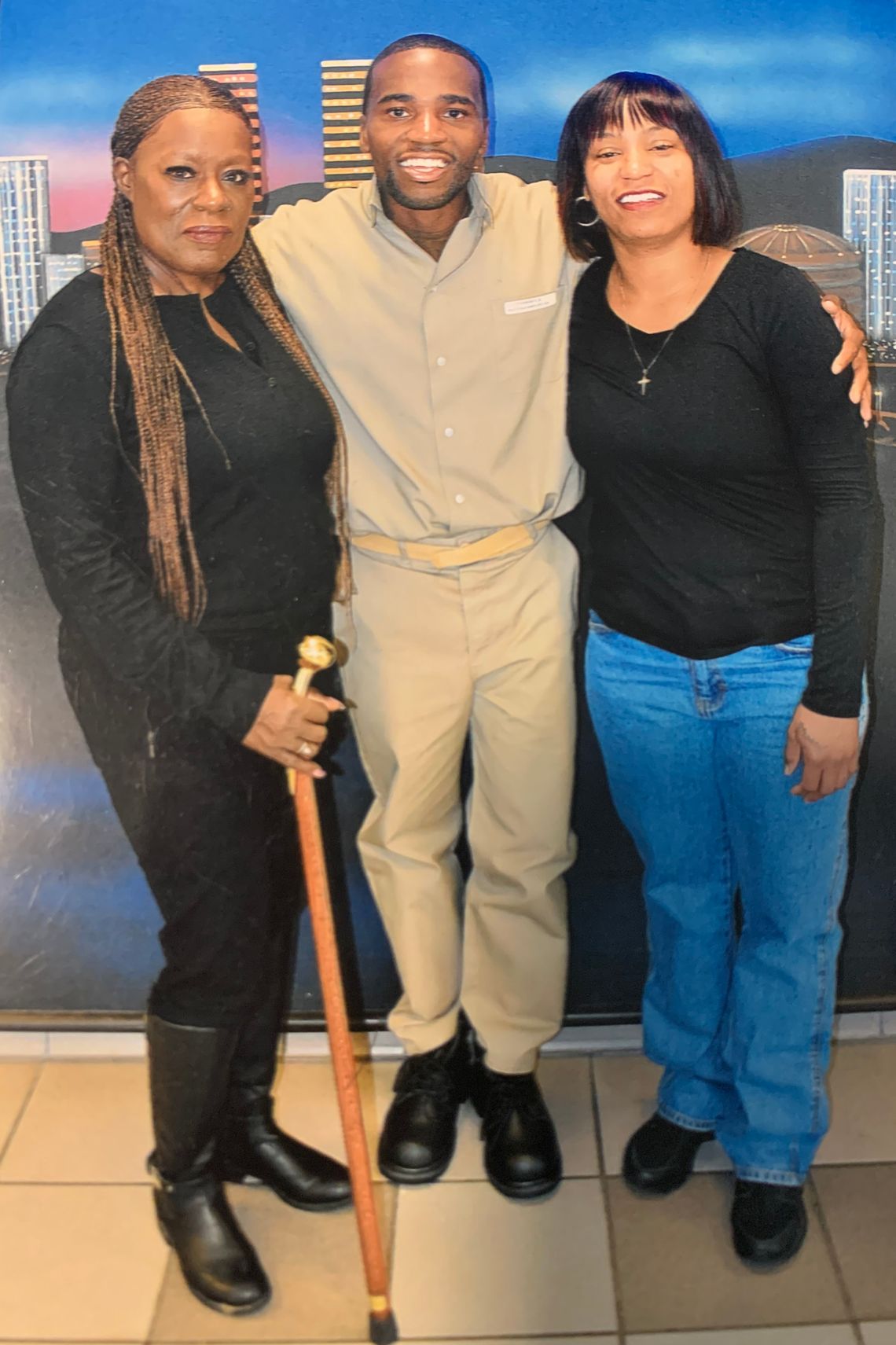 Bobby Everson stands in between his aunt Angela and his sister Ebony during a prison visit. Bobby is wearing a tan prison uniform. Angela is wearing black clothing and holds a cane. Ebony is wearing a black shirt and blue jeans. 