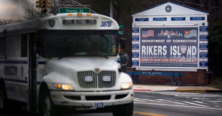 A white bus drives in front of a sign that says "Department of Correction: Rikers Island."  