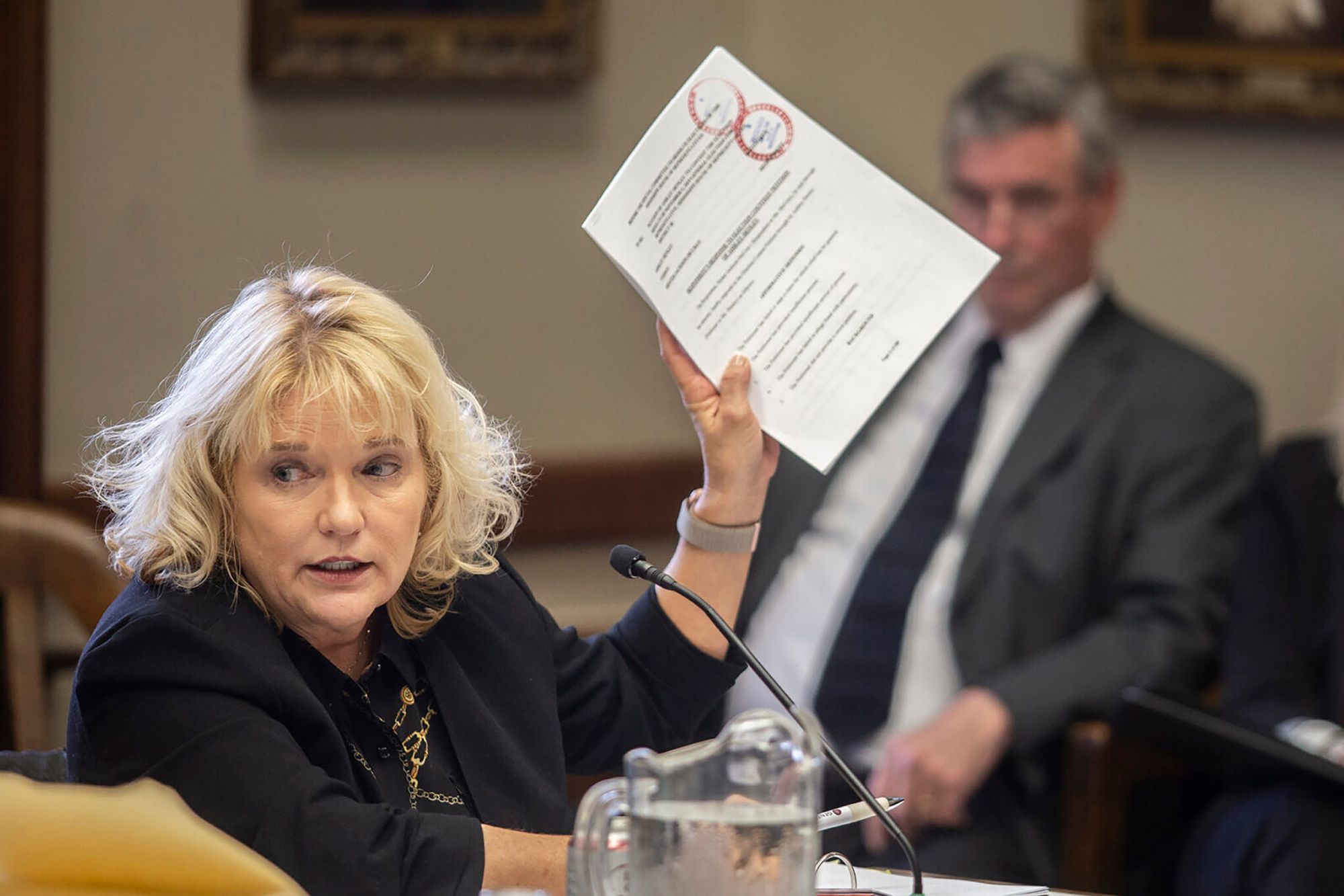 A photo of a White woman with short blonde hair in a black suit, speaking into a mic as she holds up a paper document. Two people are sitting in the background. 