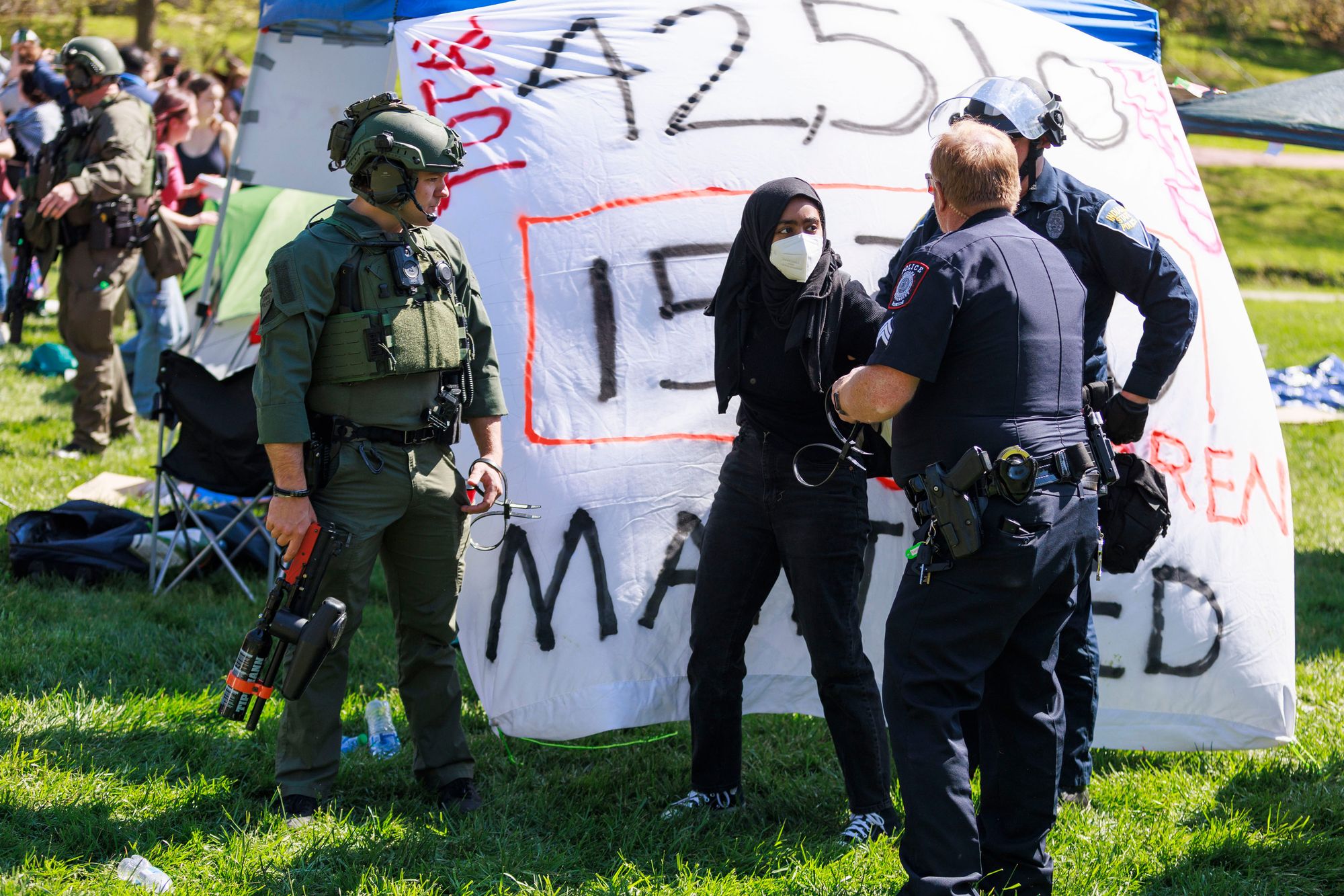 Police officers in riot gear arrest a person wearing a hijab and a mask. 