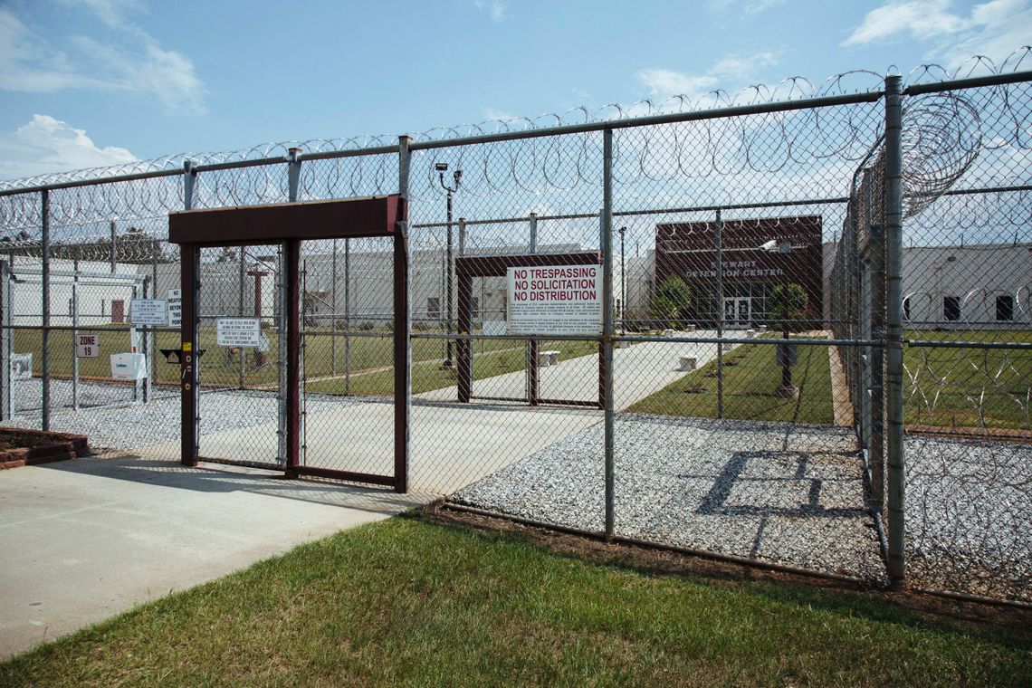 A chain-link fence topped with barbed wire is in the foreground, with a chain-link door outlined in dark red. A sign on the fence to the right reads, "No Trespassing, No Solicitation, No Distribution." Another chain-link fence with a gate door sits past the first fence, and a path leads to a building that has words on it saying, "Stewart Detention Center."