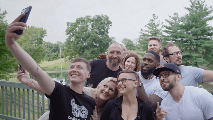 A group of people, including formerly incarcerated people, pose for a selfie with one of them holding out their phone, on a deck that overlooks a small pond nestled among some green trees.