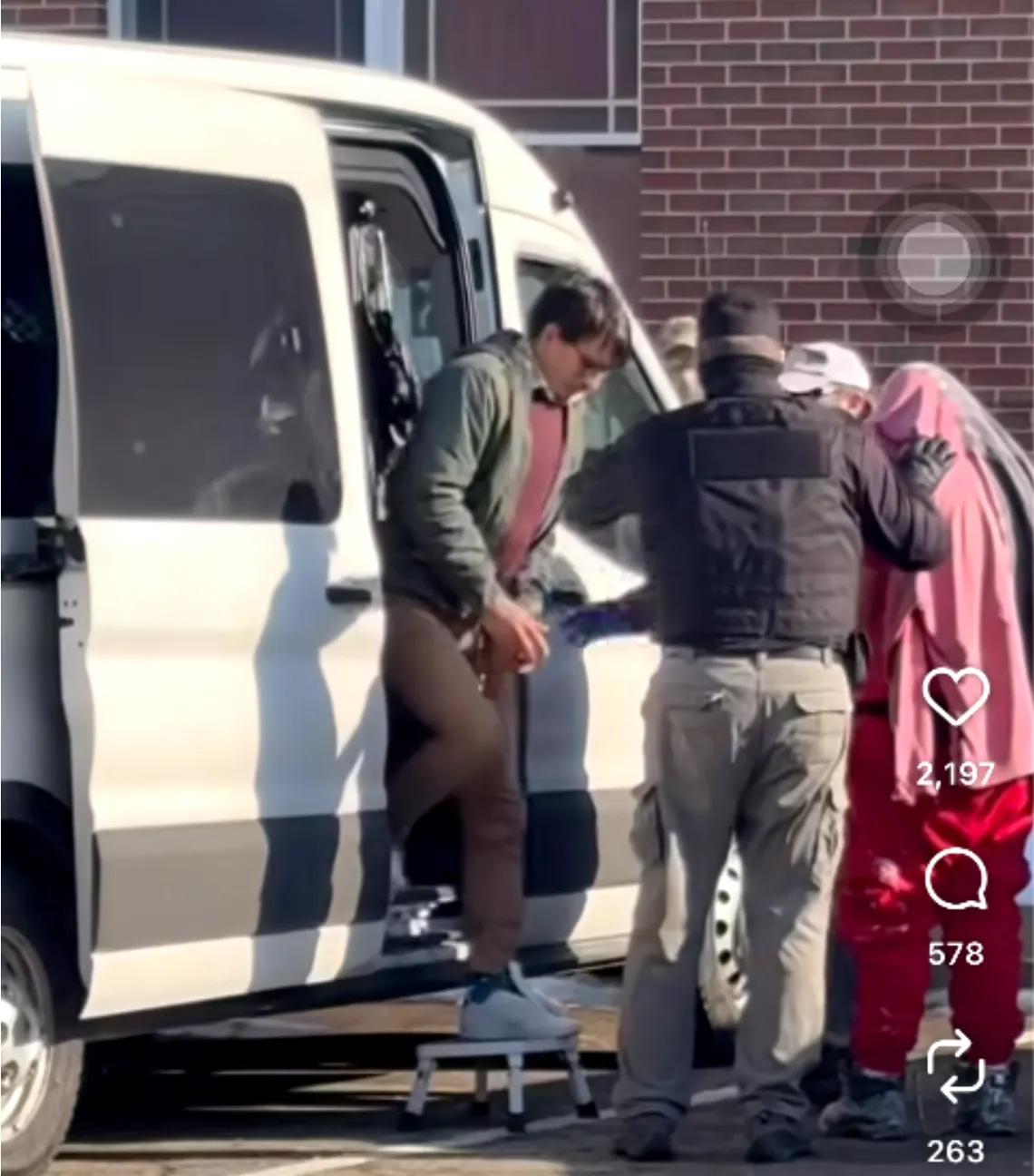 A photo shows a man with dark brown hair being led out of a white van by another person wearing a dark top and khaki pants. A person to the right has their head covered by a garment or fabric, and they are wearing red pants. 