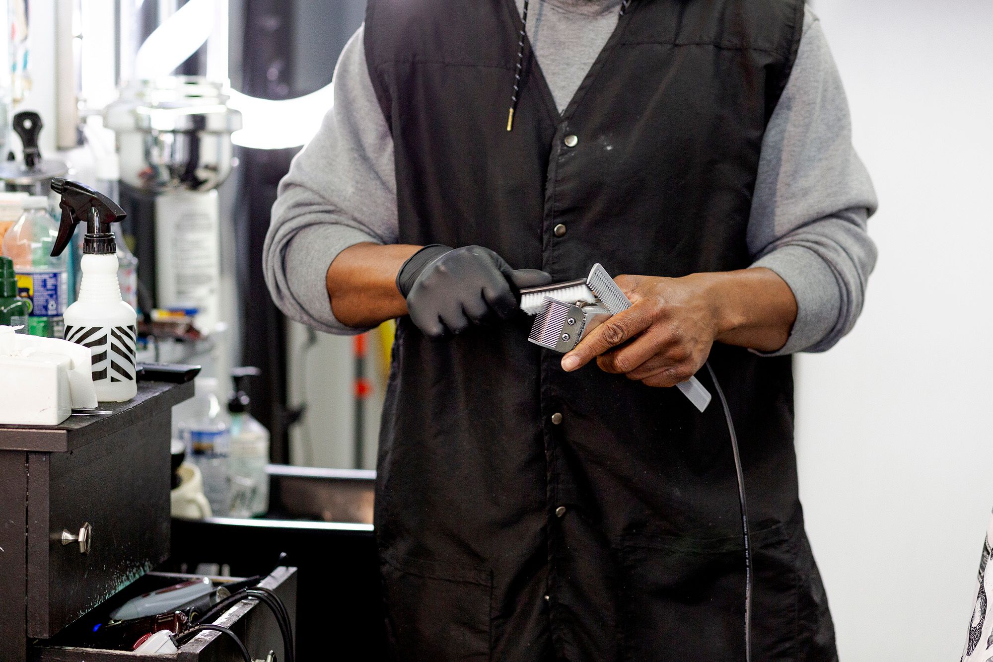 A Black man, in a black vest and gray sweater, brushes a hair clipper. 