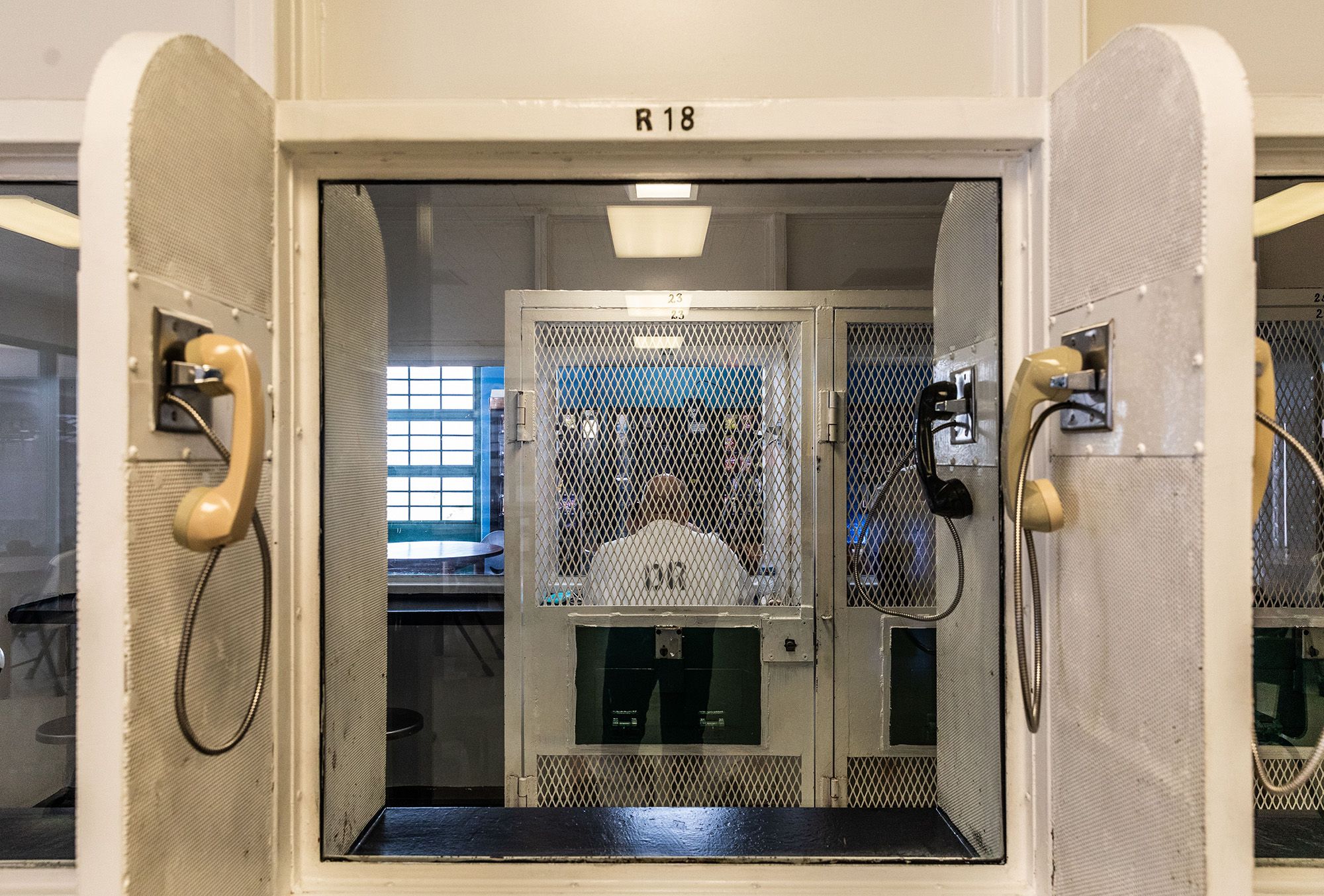 A window shows the visiting room of a prison unit, with two yellow phones hanging from the left and right wall dividers. 