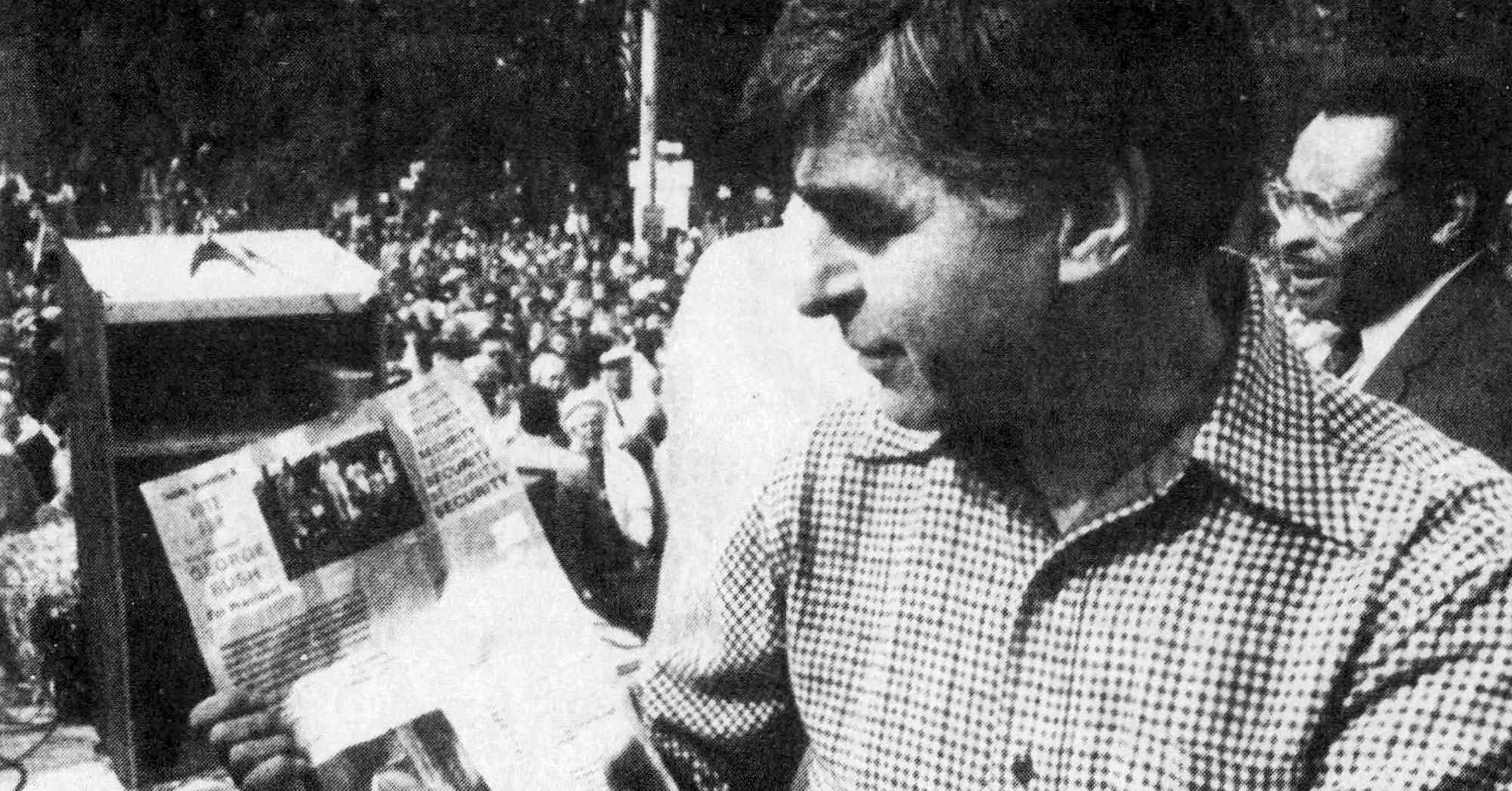 Gov. Michael Dukakis, a White man in a checkered shirt, holds a leaflet that has the words "Vote for George Bush" and "Security" printed on them, among other text that is not visible.  