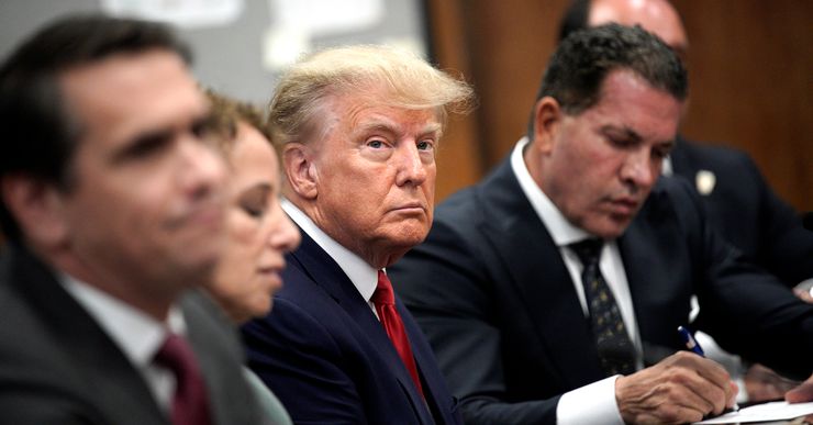 A White man, in a red tie and navy suit, sits in between his four attorneys in a courtroom.