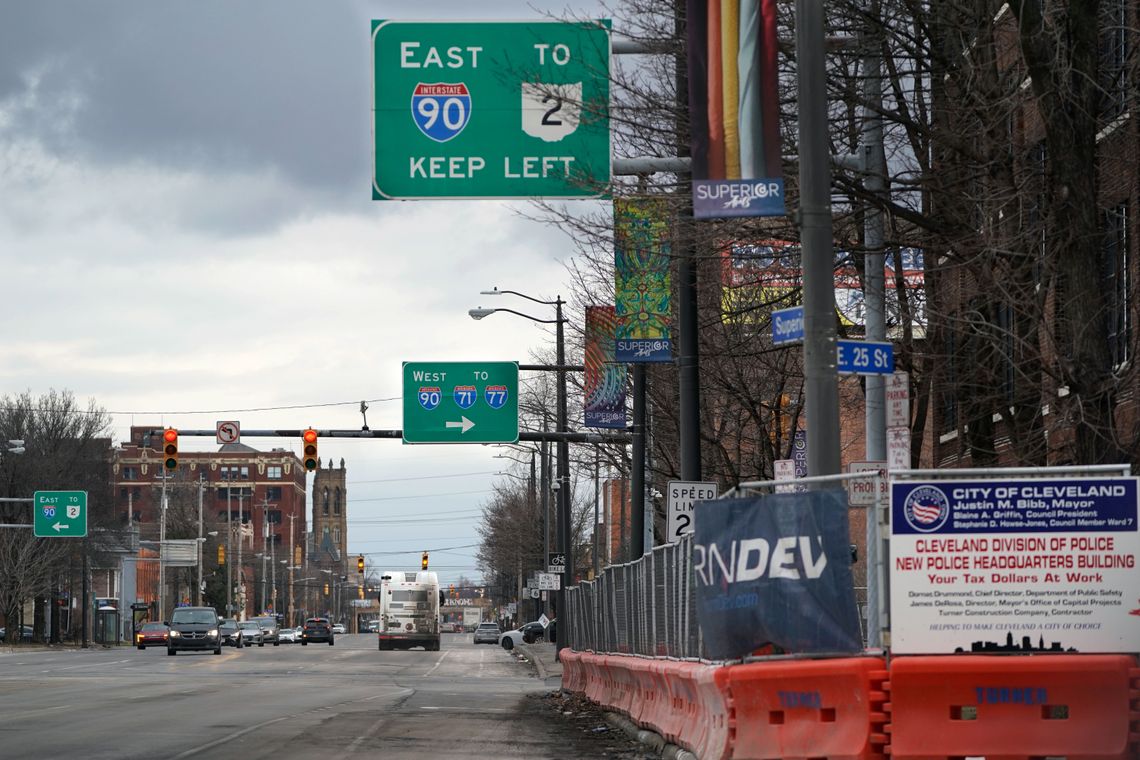 Signs for Interstate 90 hang above a street with cars on it. On the right is a fence with a sign that says “Cleveland Division of Police, New Police Headquarters Building.” 
