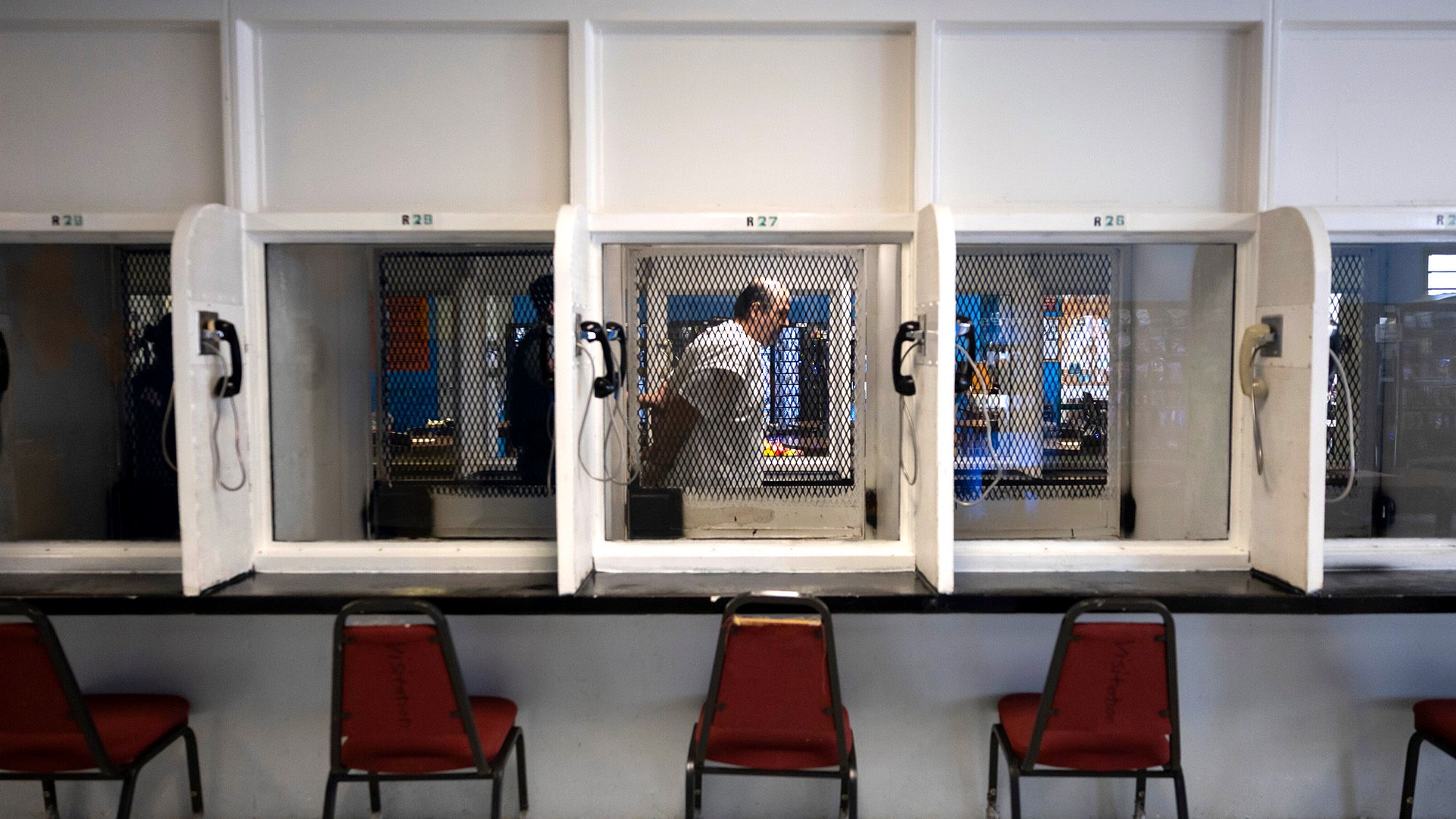 A photo shows a White man, in a white prison uniform with his hands behind his back, standing behind visitation room glass. 