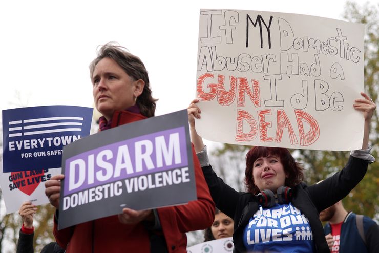 A woman, wearing a blue "March for Our Lives" shirt and black blazer, holds a sign that reads, "If my domestic abuser had a gun, I'd be dead." Standing to her left is a person holding a sign that reads, "Disarm domestic violence." 