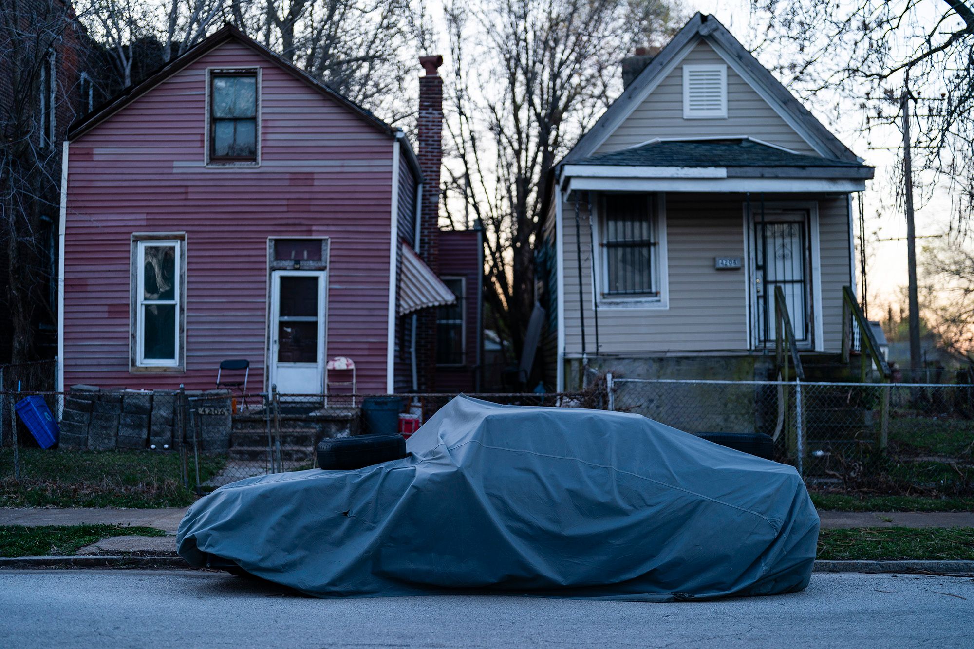 A photo shows a tarp-covered car parked on a street in front of a red house and a tan house. 