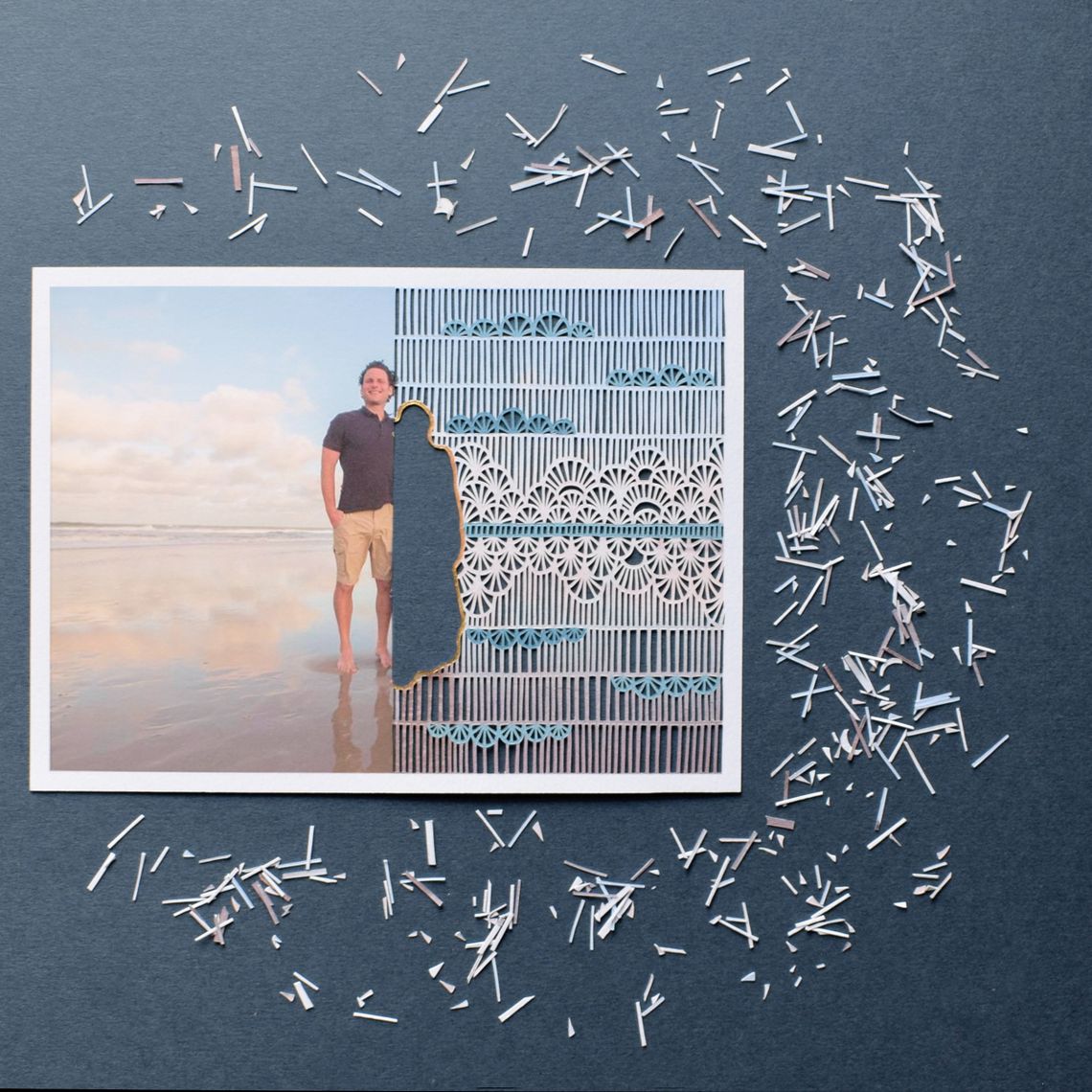 Leon Jacob, a White man wearing a dark shirt and khaki shorts, stands at the beach in a photo, with the shape of a woman cut out and leaning next to him. The half of the photo that has the woman’s shape cut out has an intricate patterned background cut out behind her. Tiny pieces of cut paper are scattered around the photo. 