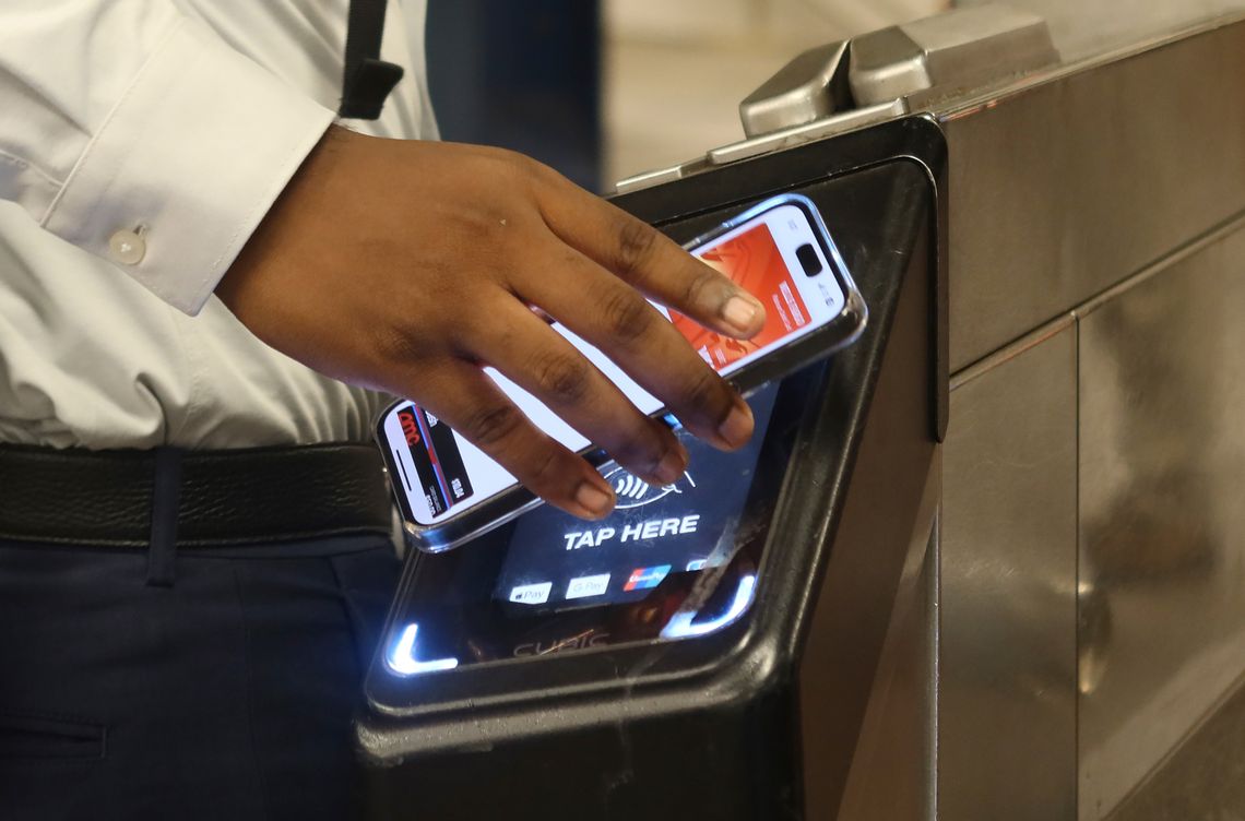 A photograph shows the hand of a person with a medium-dark skin tone using a smartphone to tap on a subway turnstile. 